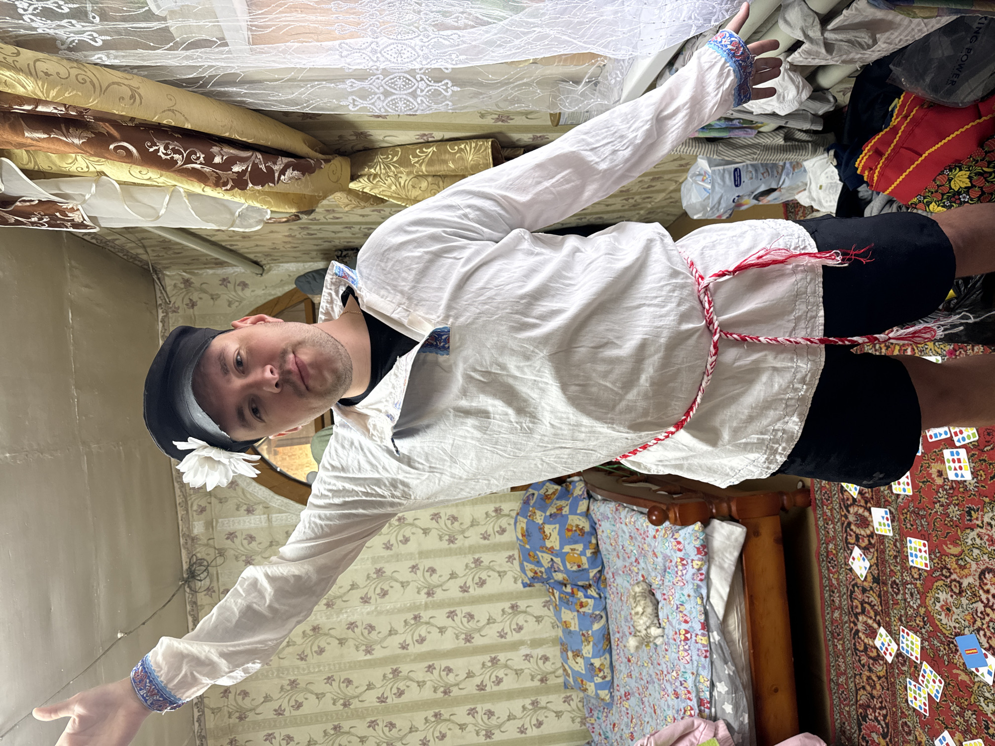 A person wearing a white shirt, black shorts, and a black cap with a white flower stands in a cluttered room in Unknown, Unknown. The person has their arms outstretched to the sides. A red and white rope-like item is tied around their waist. The room contains a bed with blue and floral patterned bedding, a wooden headboard, and a rug with scattered playing cards. The wallpaper has a floral pattern. A pile of various items, including clothing and packaging, is in the background. Curtains and drapes are hanging in the upper left corner.