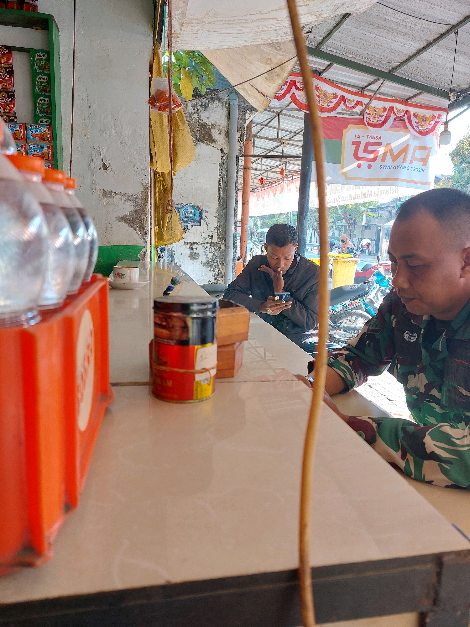 The image depicts a roadside stall in Sumuragung, Indonesia. In the foreground, an orange plastic container holds several clear plastic bottles with orange caps. To the right, a man in camouflage uniform is seated at a counter, looking down. Further back, another man wearing a dark jacket is also seated, looking at his phone. Behind them, a banner with red and white Indonesian flags and the text "LA-TANSA SMA SWALAYAN & GROSIR" is visible, suggesting the presence of a supermarket or wholesale store. The environment appears to be outdoors, with a roof overhead and a street scene with motorcycles and people visible in the background. The overall atmosphere suggests a casual, everyday setting at a local business.
