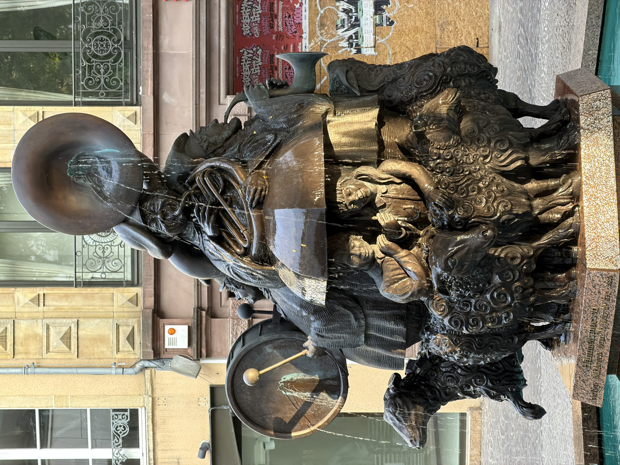 This is a detailed, matter-of-fact description of the image:
The image shows a bronze fountain sculpture situated outdoors in Luxembourg City, Luxembourg. The sculpture depicts a scene with multiple figures and animals, all rendered in dark bronze and actively involved with water.
At the top of the sculpture, a large tuba-like instrument is the source of water, which cascades down. Below this, several figures are integrated into the composition. One figure is playing a large drum with a mallet, another appears to be playing a horn or trumpet, and yet another is holding a musical instrument that resembles a trombone or french horn. These figures are dressed in what appears to be period clothing with hats.
Underneath these figures, a large, dark umbrella-like structure is positioned. Beneath this umbrella, two smaller figures, possibly children, are seated. One child is looking forward with hands clasped, and the other is also looking forward.
At the base of the sculpture, several sheep are depicted. Their wool is intricately textured with swirling patterns. One sheep is to the left, with its head turned and water dripping from its muzzle. Other sheep are clustered together, forming the lower part of the fountain's structure. Water flows over and around the sheep and into a pool below.
The sculpture is mounted on a multi-tiered base. The bottom tier is a rough-textured pink granite block with an inscription that is partially visible and appears to read "HEMMELSGESELLSCHAFT voriger Völkerschau". The base rests in a shallow, blue-tiled pool, indicating it is an active water feature.
In the background, there is a building with yellow and beige facade. It features architectural details like recessed panels and decorative window grills. A grey downspout runs vertically down the building. There are also posters affixed to the building in the background, some with red backgrounds and graffiti-like text, suggesting a public or commercial area. The lighting indicates it is daytime, with shadows cast by the sculpture.
