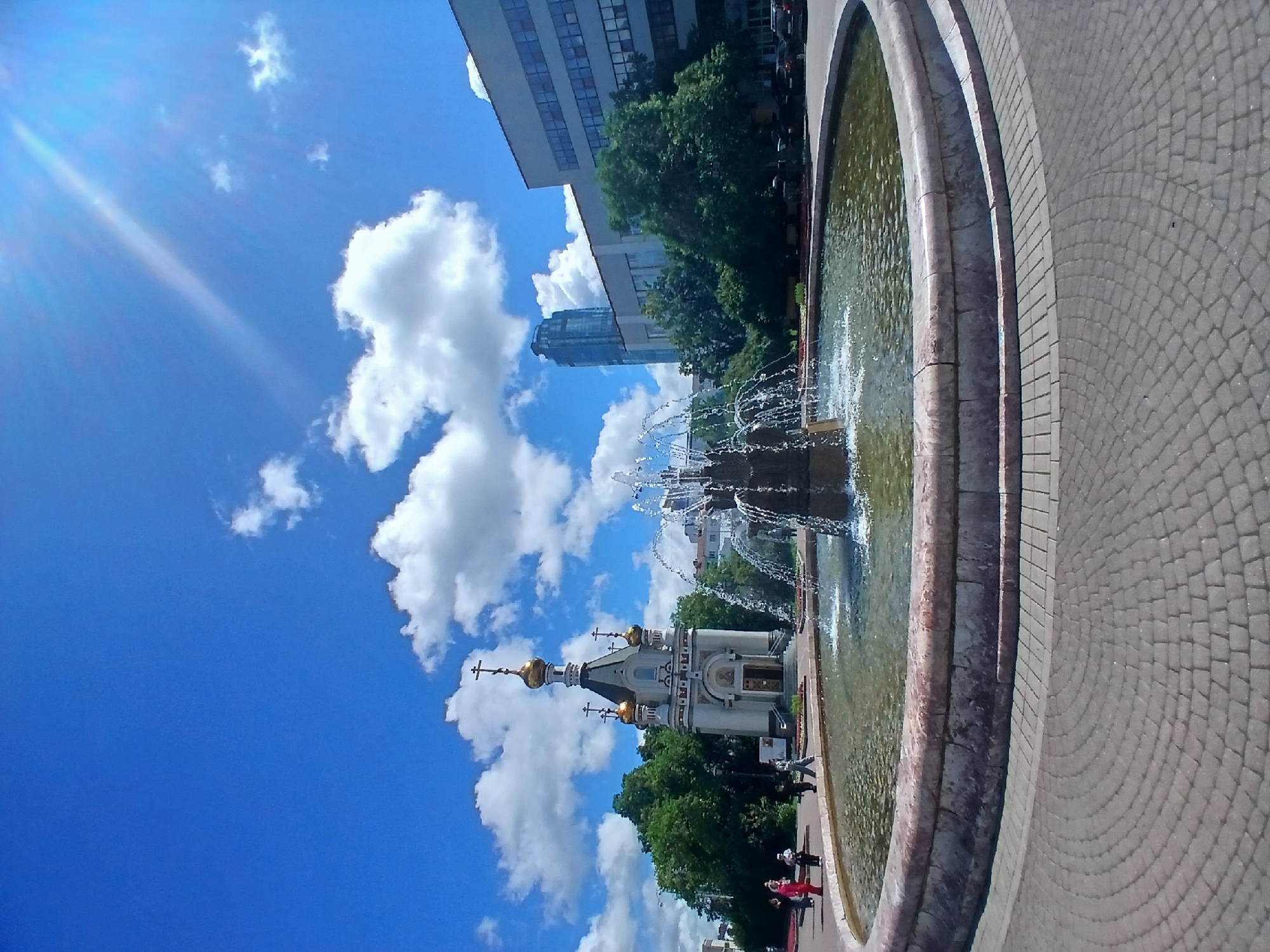 This media file captures an outdoor scene featuring a fountain in the foreground. The fountain is circular with a stone rim and contains water with visible jets. Adjacent to the fountain is a cobblestone pathway. In the middle ground, a small, ornate church with golden domes is visible, surrounded by lush green trees. Beyond the trees, modern buildings, including a tall glass skyscraper, are present. The sky is a vibrant blue with scattered white clouds, and sunlight streams into the frame, suggesting a bright, clear day. A few people are seen walking along the pathway near the church and fountain. The image appears to be taken from a slightly elevated angle, looking down and across the scene. The orientation of the image is vertical. The overall impression is of a pleasant, sunny day in an urban environment that blends historical and modern architecture with natural elements.