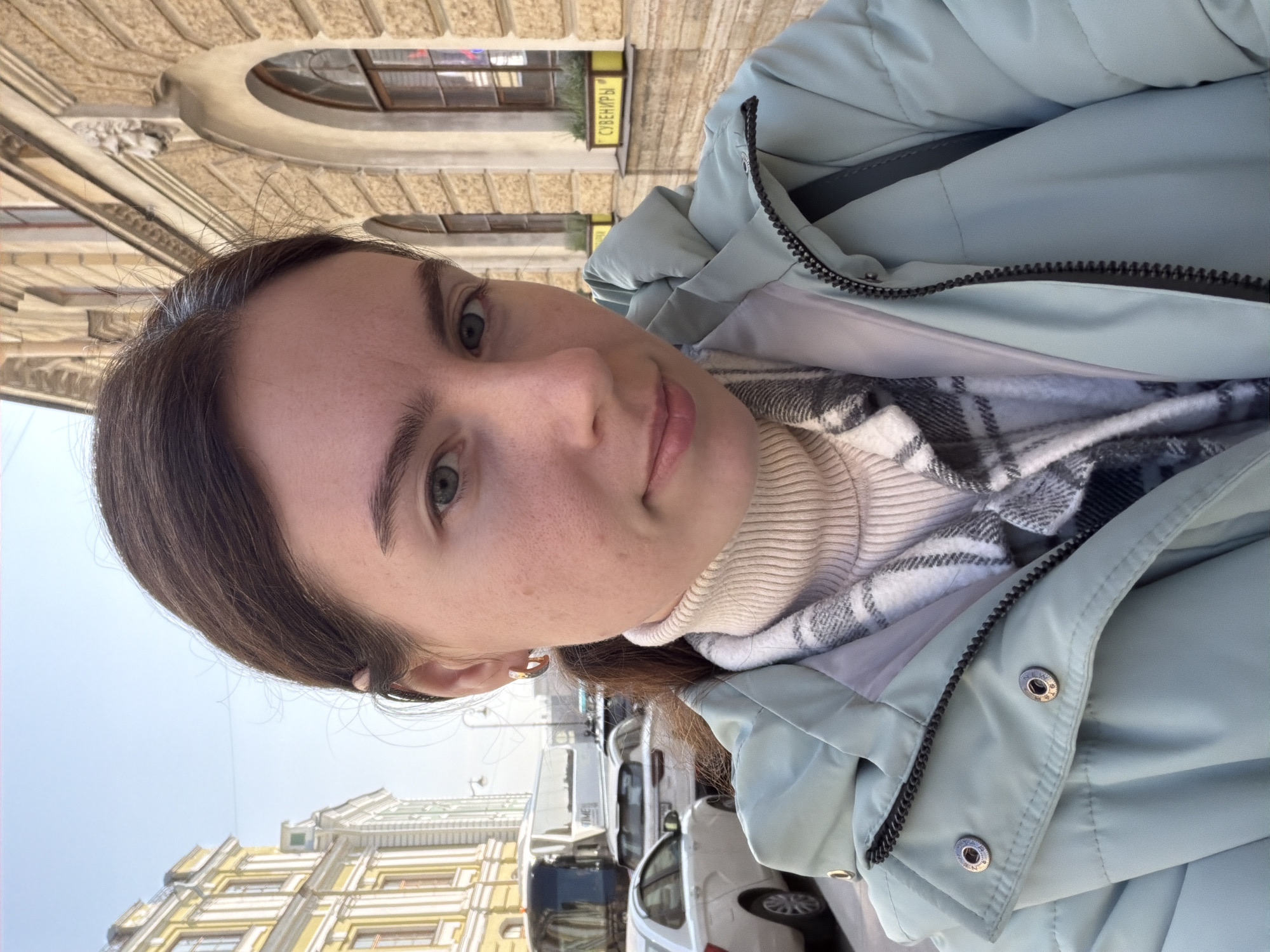 A woman with brown hair and fair skin is taking a selfie outdoors. She is wearing a light blue puffer jacket, a white turtleneck sweater, and a gray and white plaid scarf. She has an earring in her left ear. The background reveals an urban environment, with buildings exhibiting classical architecture, some with yellow facades. Cars are parked along the street. A sign with Cyrillic text, "СУВЕНИРЫ," is visible on one of the buildings, indicating the location is likely in Russia. Given the architectural style, the city is likely Saint Petersburg.