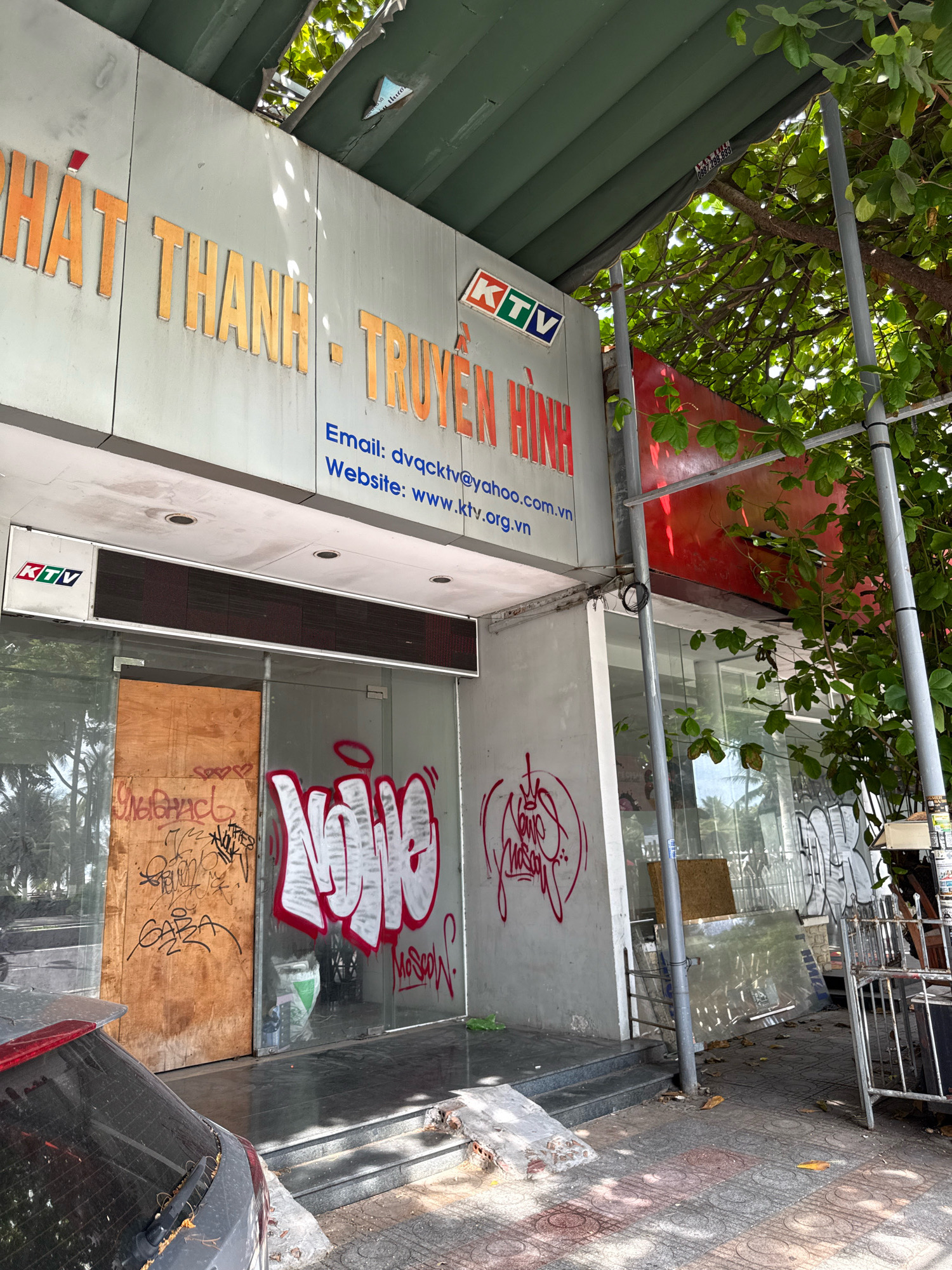 The image shows the exterior of a building in Nha Trang, Vietnam, with graffiti on the windows and a boarded-up entrance. The building appears to be an abandoned or closed business, as indicated by the graffiti and boarded-up entrance. The signage suggests it was a media company, with the text "PHÁT THANH - TRUYỀN HÌNH" (Broadcasting - Television) and a KTV logo visible. The overall scene is one of urban decay and neglect, with overgrowth of trees and debris on the ground. The street in front of the building is paved with tiles and has some fallen leaves. A portion of a grey car is visible in the lower left corner of the image.