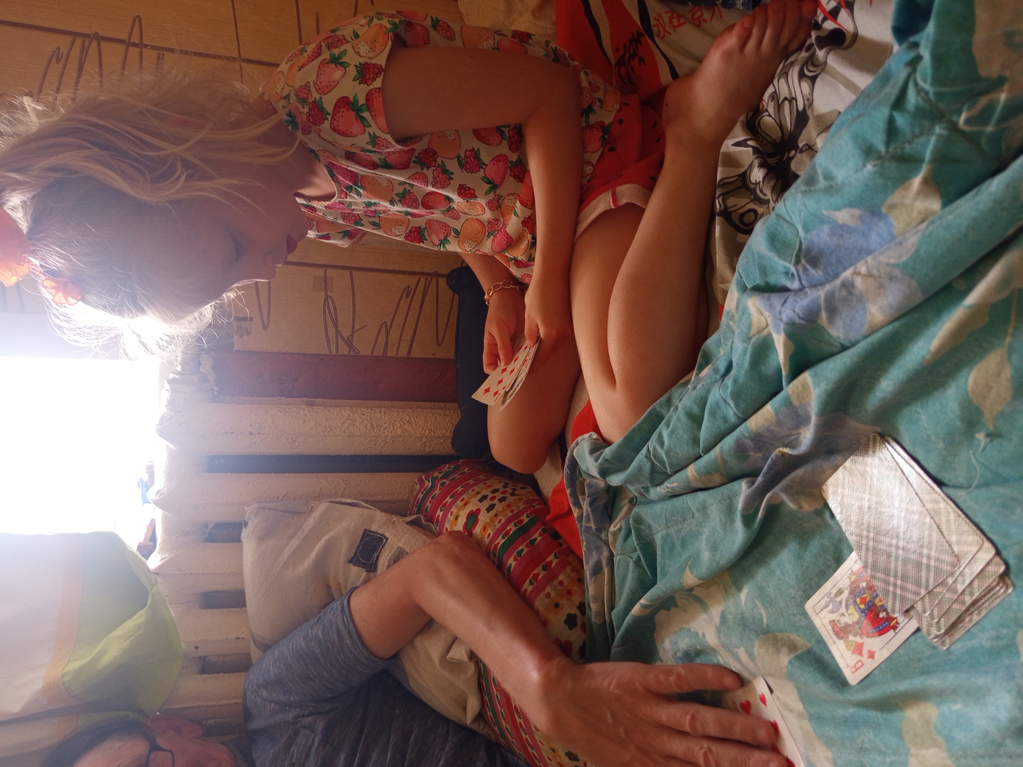 In a dimly lit room, likely in Gdov, Russia, a girl with blonde hair tied in a ponytail is engrossed in a game of cards. She wears a t-shirt with a strawberry pattern and red shorts. Her right hand holds a few cards, while her left hand is near a larger stack of cards. Across from her, a person's arm, resting on pillows, is visible, suggesting a shared activity. The environment appears to be a bedroom, with a window providing natural light, casting shadows on the wall. A radiator and a patterned blanket on the bed contribute to the domestic setting. The overall mood is one of quiet concentration and leisure.