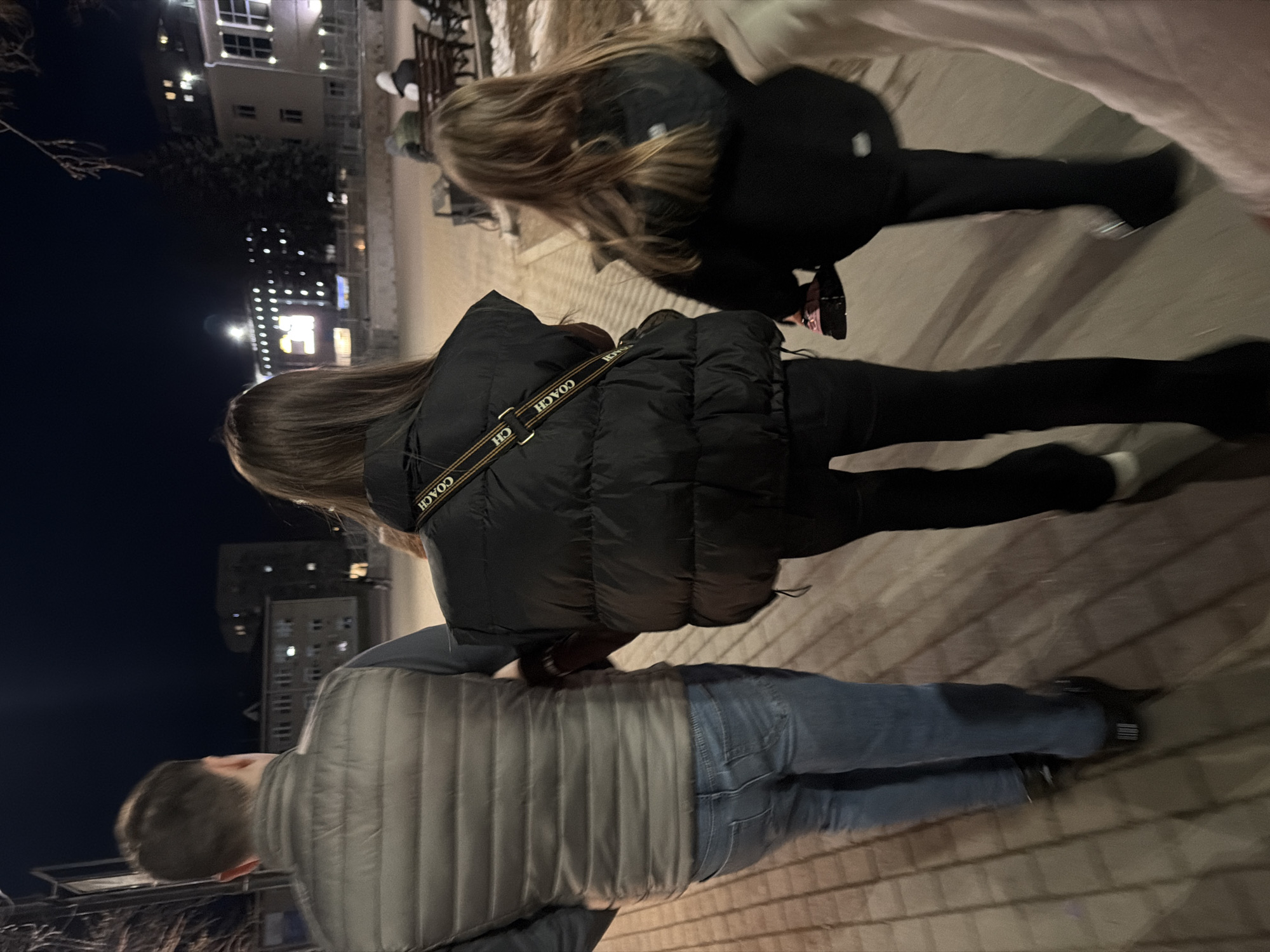 A group of three people walk on a brick walkway at night in what appears to be a city. The person closest to the camera is wearing a grey vest, blue jeans, and dark shoes. A second person is wearing a black puffer jacket with a Coach logo strap across their body, black pants, and dark shoes. A third person is wearing a black coat and black pants. Buildings with illuminated windows can be seen in the background. The location is likely Chaikovsky, Russia.