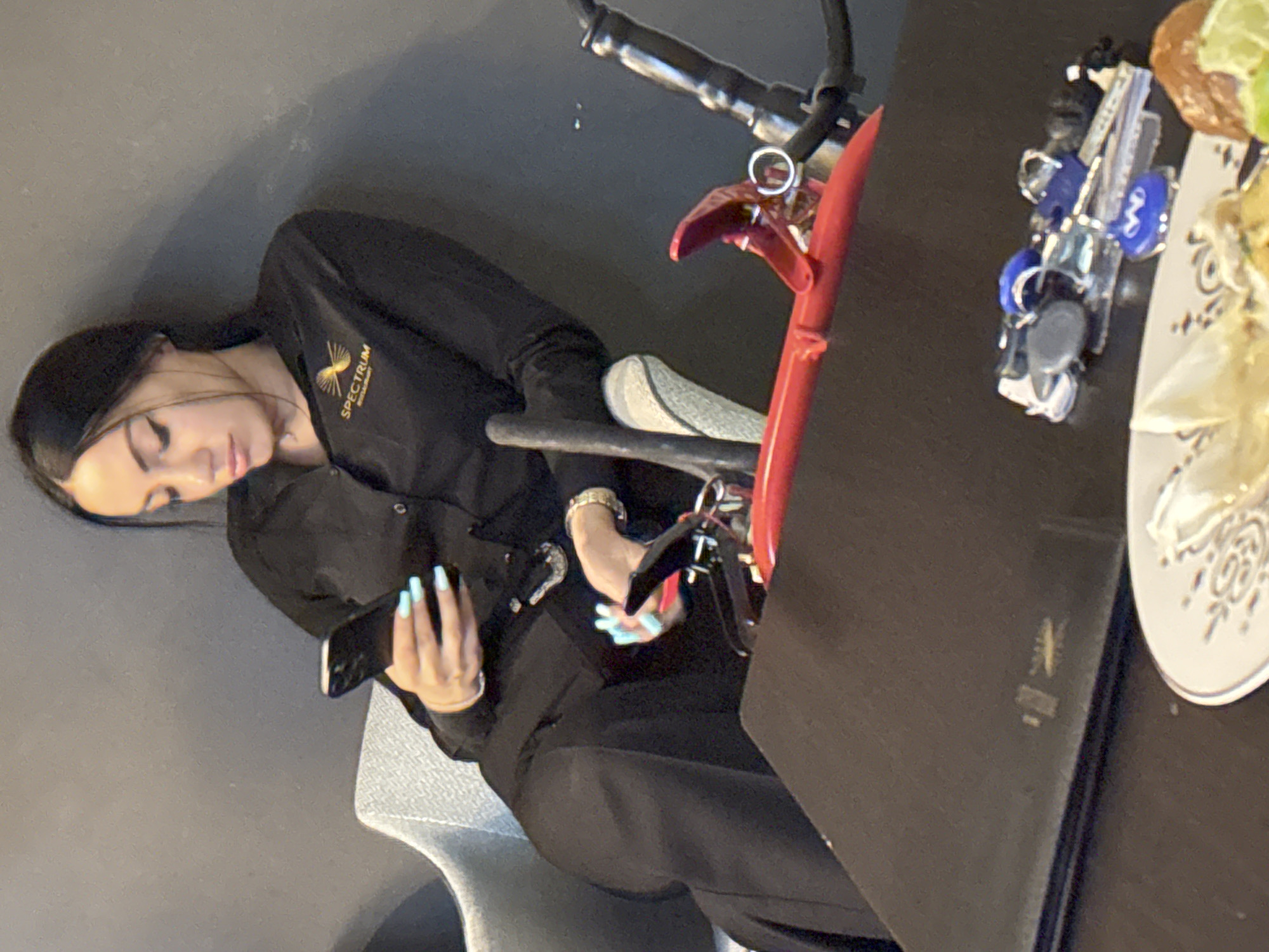 A woman with dark hair and light blue fingernails is seated in a gray chair, looking at her phone. She is wearing a black collared shirt with a gold logo that reads "SPECTRUM RESTAURANT". The background is a plain gray wall. In the foreground, a black table is visible, with a plate containing food and a set of keys on it. A red object, possibly a fire extinguisher, is positioned behind the woman. The overall scene appears to be indoors, possibly in a restaurant or lounge area. The city of Sofia, Bulgaria is not discernible from the image.