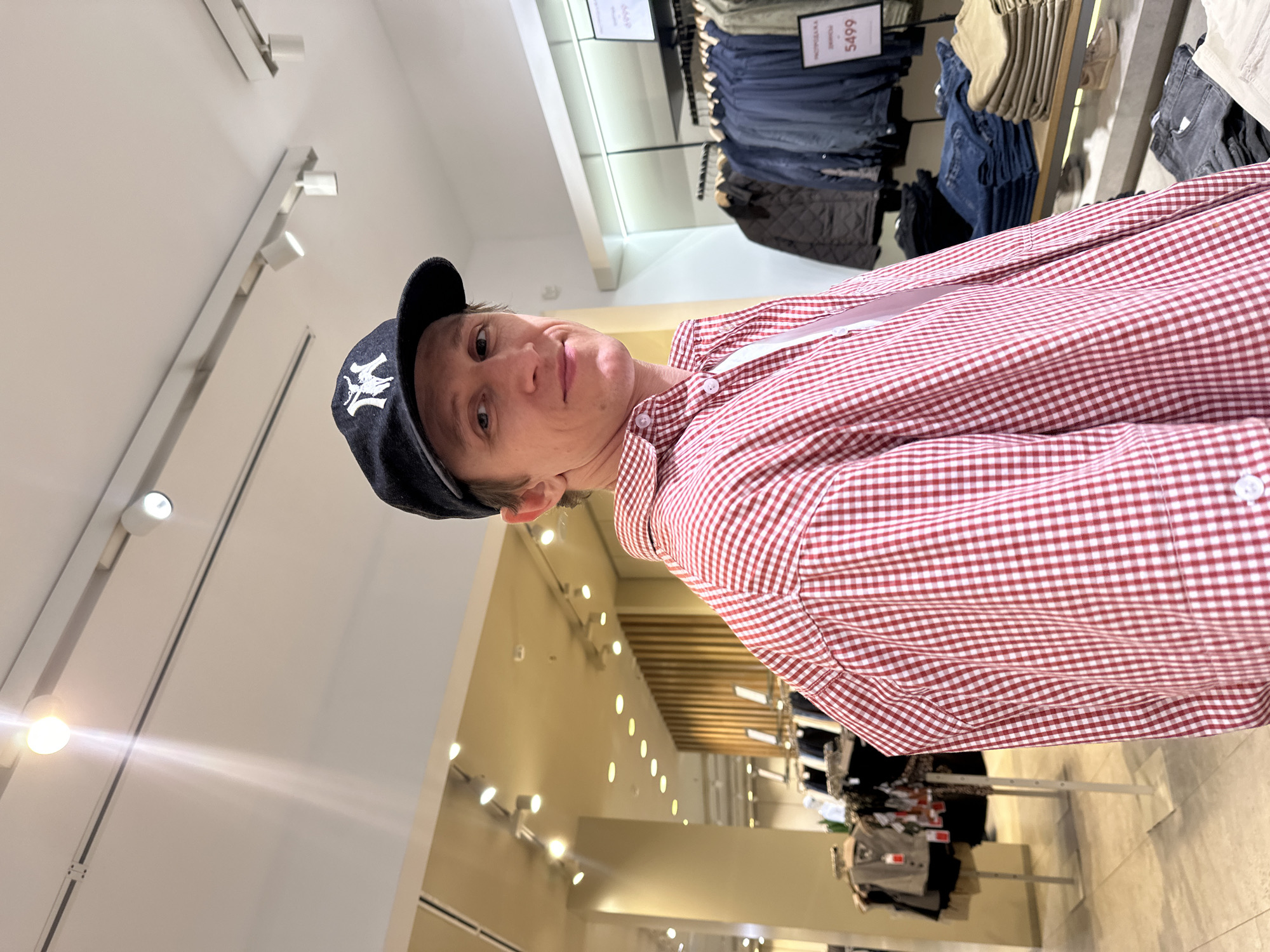A man wearing a black baseball cap with a white logo and a red and white gingham long-sleeved shirt is standing in a clothing store in Izhevsk, Russia. He is looking towards the camera. The store appears to be well-lit with track lighting on the ceiling. Clothing racks with various garments, including jackets and jeans, are visible in the background. A price tag showing "5499" is also visible on one of the racks.