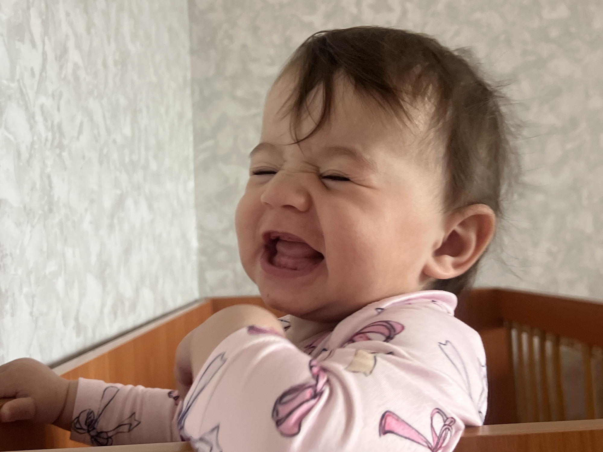 A baby with dark hair and a pink pajama with ballerina shoes is in a crib with wooden slats. The baby's face is scrunched up and their mouth is open, as if crying. The background is a light-colored wall with a textured pattern. The photo was taken in Elena, Bulgaria.