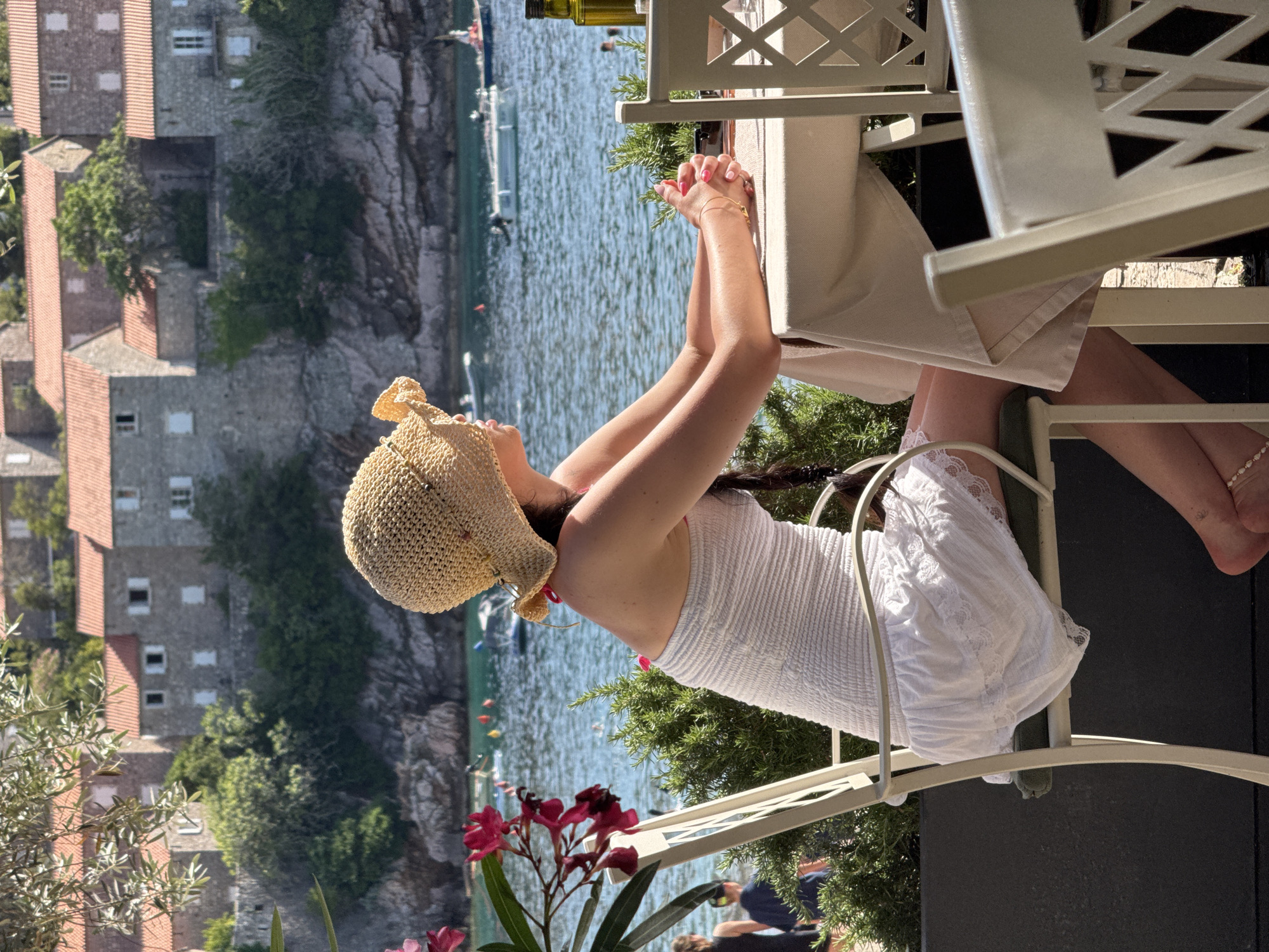 A woman, positioned in profile facing right, sits at an outdoor dining table overlooking the Adriatic Sea in Sveti Stefan, Montenegro. She wears a wide-brimmed, light-colored straw hat and a white strapless, smocked dress with a lace-trimmed skirt. Her dark hair is styled in a braid over her right shoulder. Her hands, featuring pink manicured nails, are clasped on the light-colored tablecloth. A delicate anklet is visible on her right ankle.
In the immediate foreground, to the left, vibrant pink flowers with green leaves are partially visible. Behind the woman, green bushy plants (possibly rosemary) delineate the dining area. On the table's right side, a dark green bottle, likely containing olive oil, is present. The chairs are light-colored metal, with a simple decorative lattice pattern on the backrest of the chair to the right.
The midground features calm, clear blue water with ripples, where several white boats and colorful buoys are scattered. On the left side of the water, distant figures, including a child, are visible.
The background is dominated by the distinct architecture of Sveti Stefan island: numerous multi-story stone buildings with reddish-orange tiled roofs and white-shuttered windows, densely packed on a rocky promontory. Green trees and vegetation are interspersed among the buildings and along the coastline. The sky is clear and bright, indicating a sunny day.