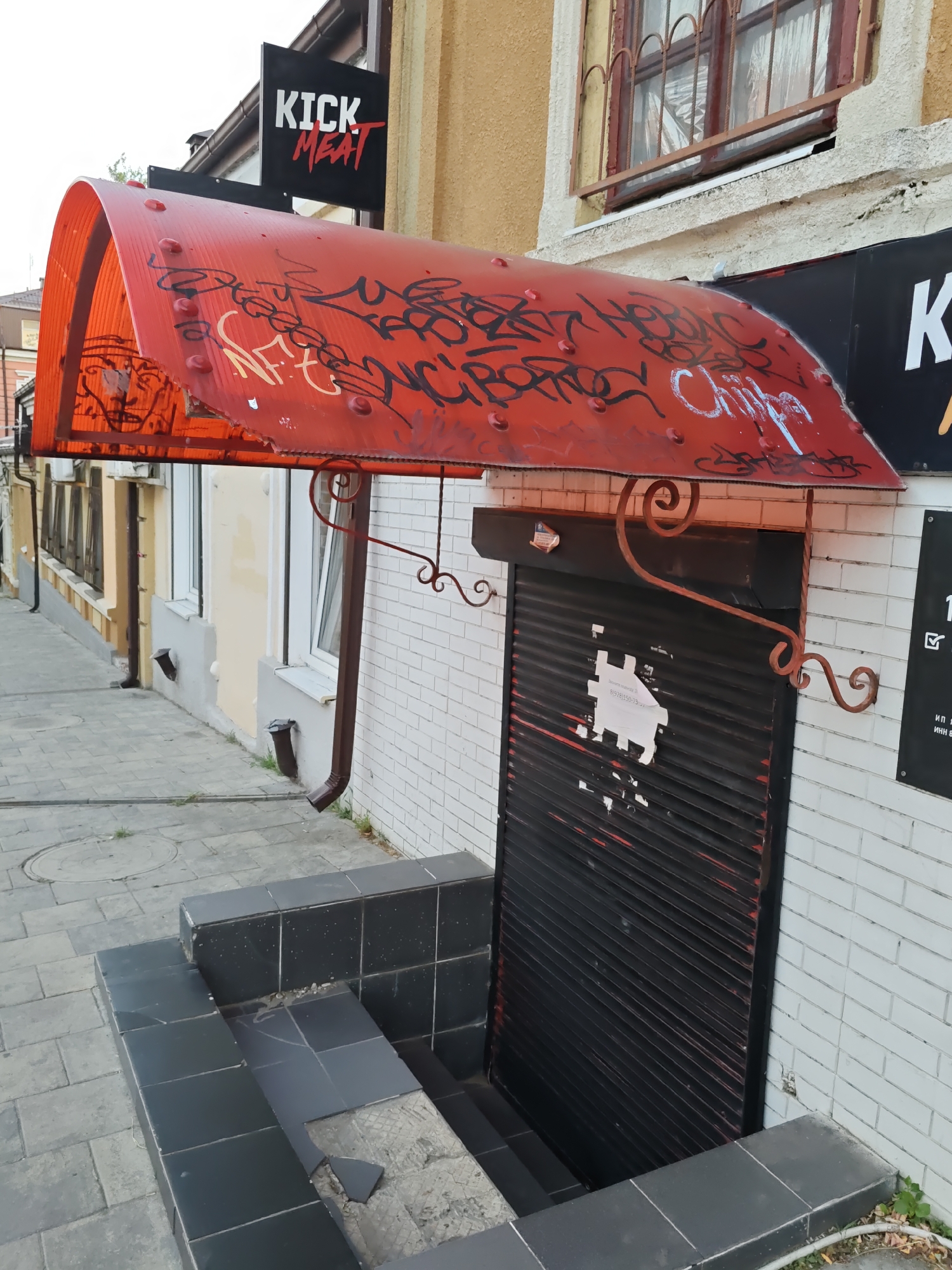 A storefront in Rostov-on-Don, Russia, features a red, corrugated plastic awning covered in black and white graffiti. Above the awning, a black sign with white lettering reads "KICK MEAT". The entrance to the establishment is a black metal roll-up door, partially open, revealing a set of descending stairs. A white paper notice is affixed to the door. The building's facade is constructed of white brick, and a yellow-painted upper story is visible. To the left of the entrance, a sidewalk made of gray paving stones leads away from the building. Black tiled steps descend to the entrance. The scene appears to be outdoors during daylight hours.
