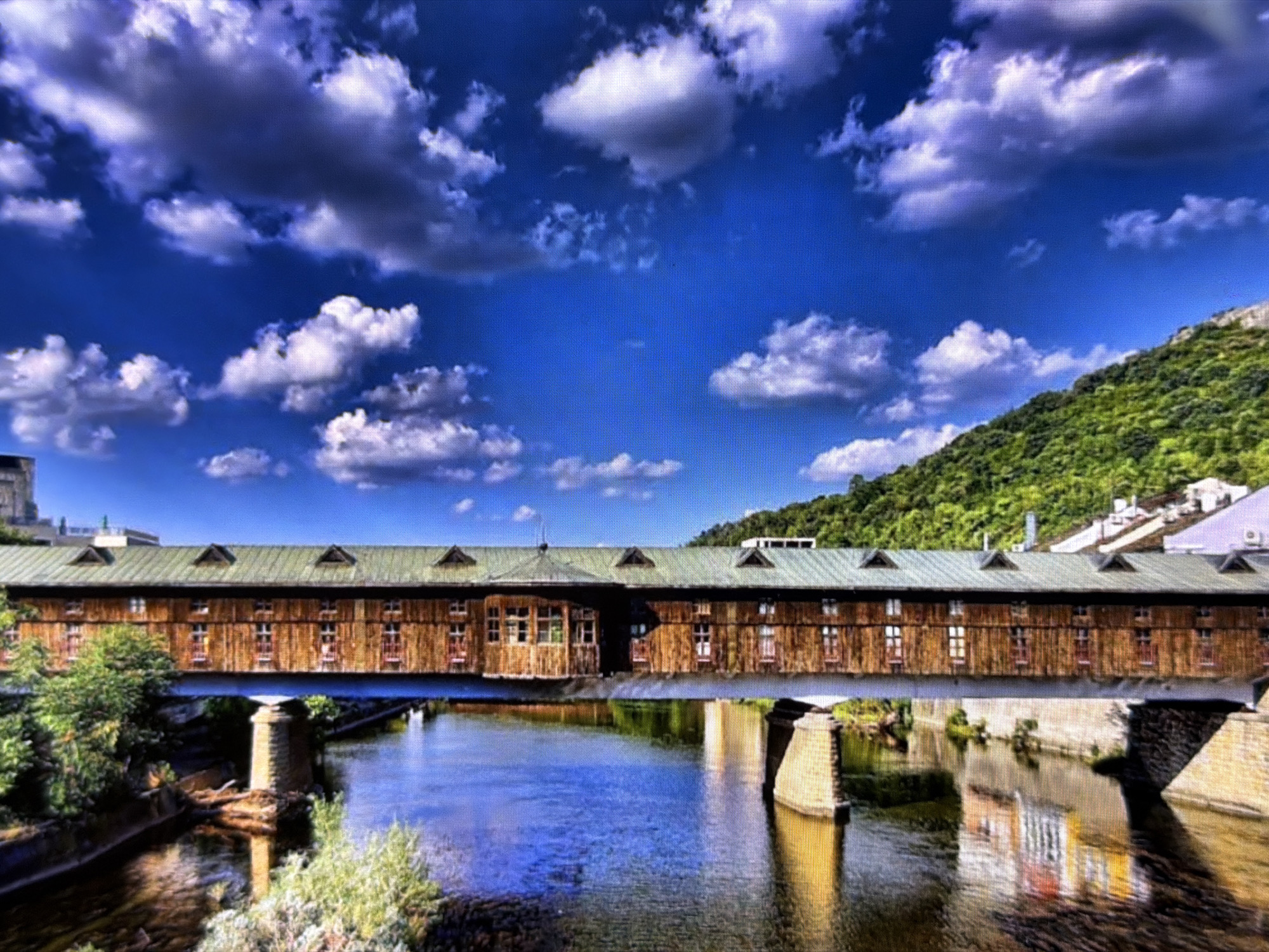 The image presents a scenic view of a long, covered wooden bridge spanning a river under a vibrant blue sky filled with scattered white and grey clouds. This is the Covered Bridge in Lovech, Bulgaria.
The bridge, the central subject, is constructed from warm brown timber with a distinctive green gabled roof featuring several small dormer windows. Its facade is adorned with numerous rectangular windows, some appearing like small doors, all framed in a dark color. The structure is supported by sturdy stone pillars embedded in the river.
On the left bank of the river, lush green trees and vegetation are visible, while on the right side, a steep, forested green hill rises in the background. Some white buildings can be seen faintly nestled against the hillside beyond the bridge. The river below reflects the bright blue sky and elements of the bridge, creating a sense of depth and mirroring the overhead light. The water appears calm with subtle reflections.
The scene is captured during the daytime, likely under bright sunlight, which illuminates the textures of the wooden bridge and the natural landscape. No people are visible, and there is no discernible text in the image. The overall impression is one of tranquility and natural beauty, highlighting a significant architectural landmark.