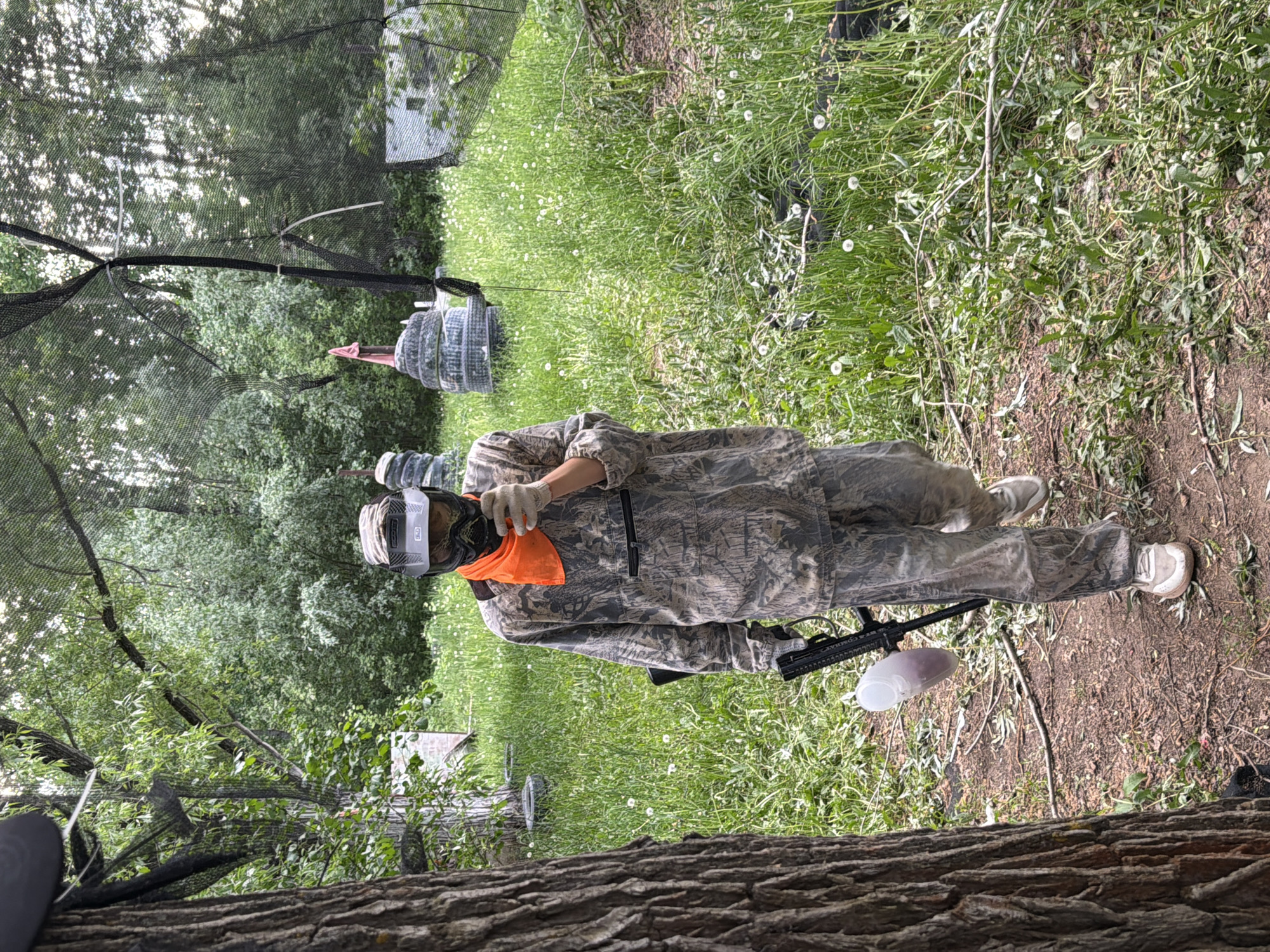 A person in camouflage clothing stands in a grassy outdoor area, holding a paintball marker. They are wearing a protective mask, gloves, and an orange cloth around their neck. The ground is covered with grass, weeds, and some dirt patches. A thick tree trunk is visible at the bottom of the frame, and netting is visible above. In the background, there are trees and some paintball field structures, including inflatable bunkers. The location may be in Chaikovsky, Russia.