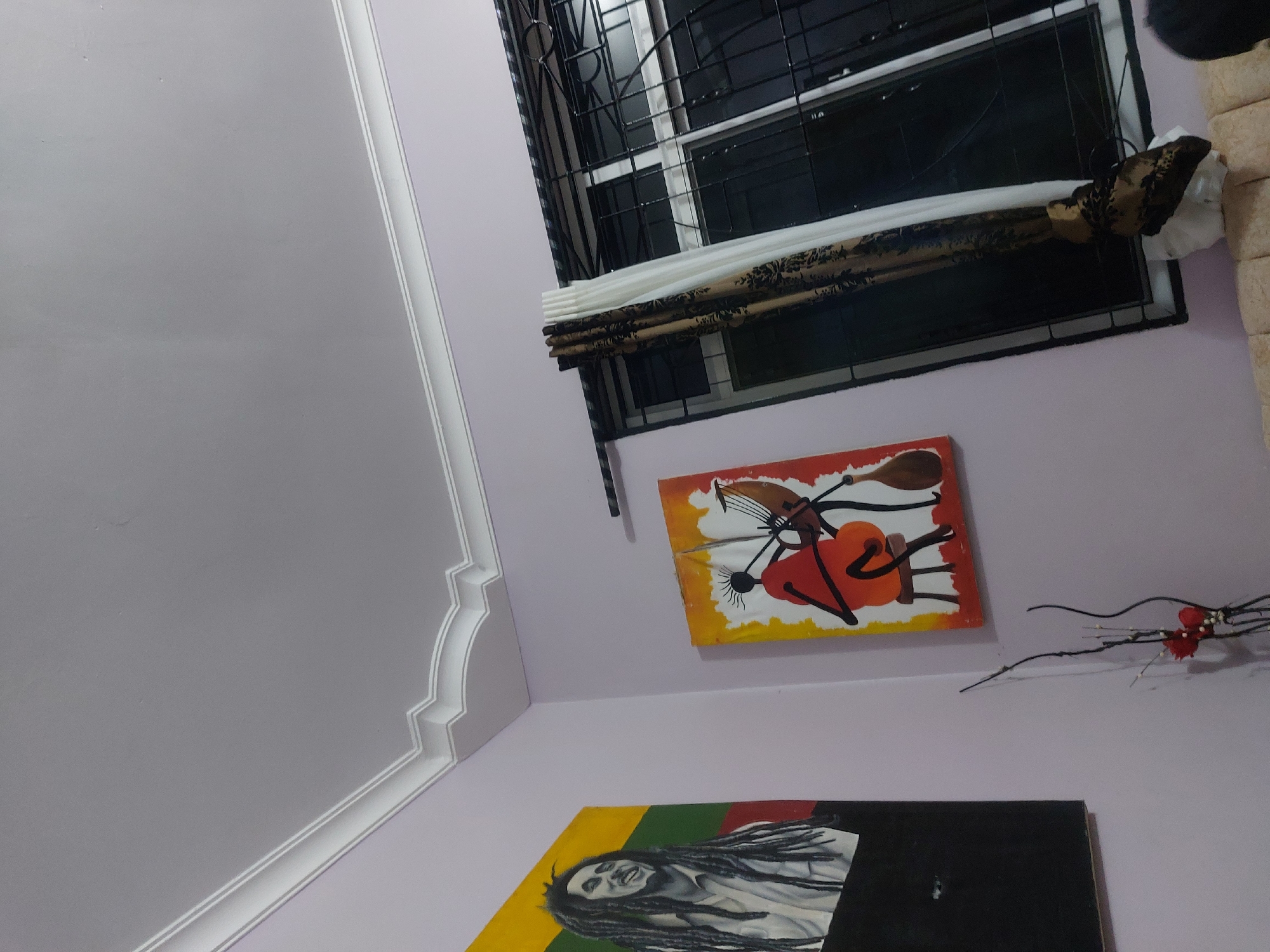 The image presents an interior scene, likely a room in a residence in Dar es-Salaam, Tanzania, as indicated by the artwork and architectural details. The walls are painted a light lavender color, and the ceiling is white with decorative molding. A window with a black metal security grill is visible, covered by a partially drawn patterned blind. Beneath the window, two pieces of artwork hang on the wall. The top artwork is abstract and features orange, yellow, and white colors. Below that is an artwork depicting Bob Marley in yellow, green, white and black. To the right of the top artwork, there is a decorative twig arrangement with small red accents.