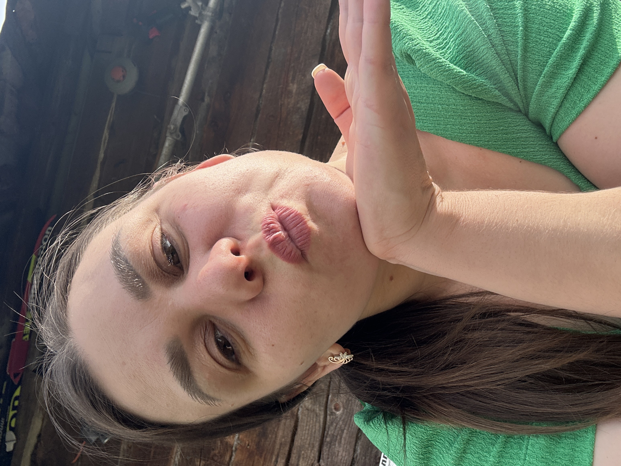 A woman with brown hair and wearing a green shirt poses for a selfie. She is pursing her lips and holding her hand near her face. She is outdoors with a wooden structure behind her. The location is Chaikovsky, Russia.