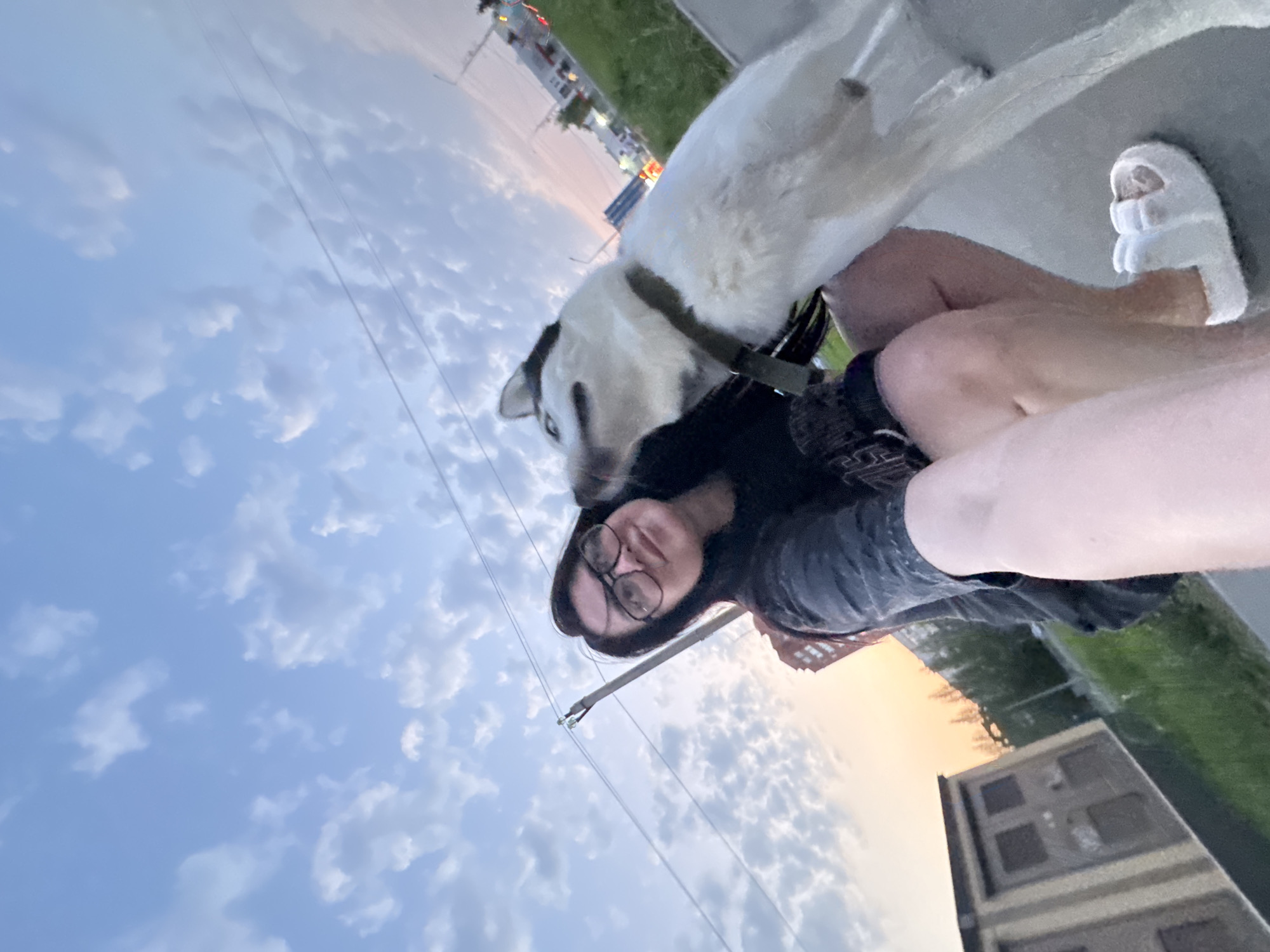 A woman wearing glasses lies on the ground with a white and black dog near a sidewalk in a city. The sky is visible with clouds. The woman wears a dark t-shirt and fuzzy slippers. The dog wears a collar. A building and power lines are in the background.