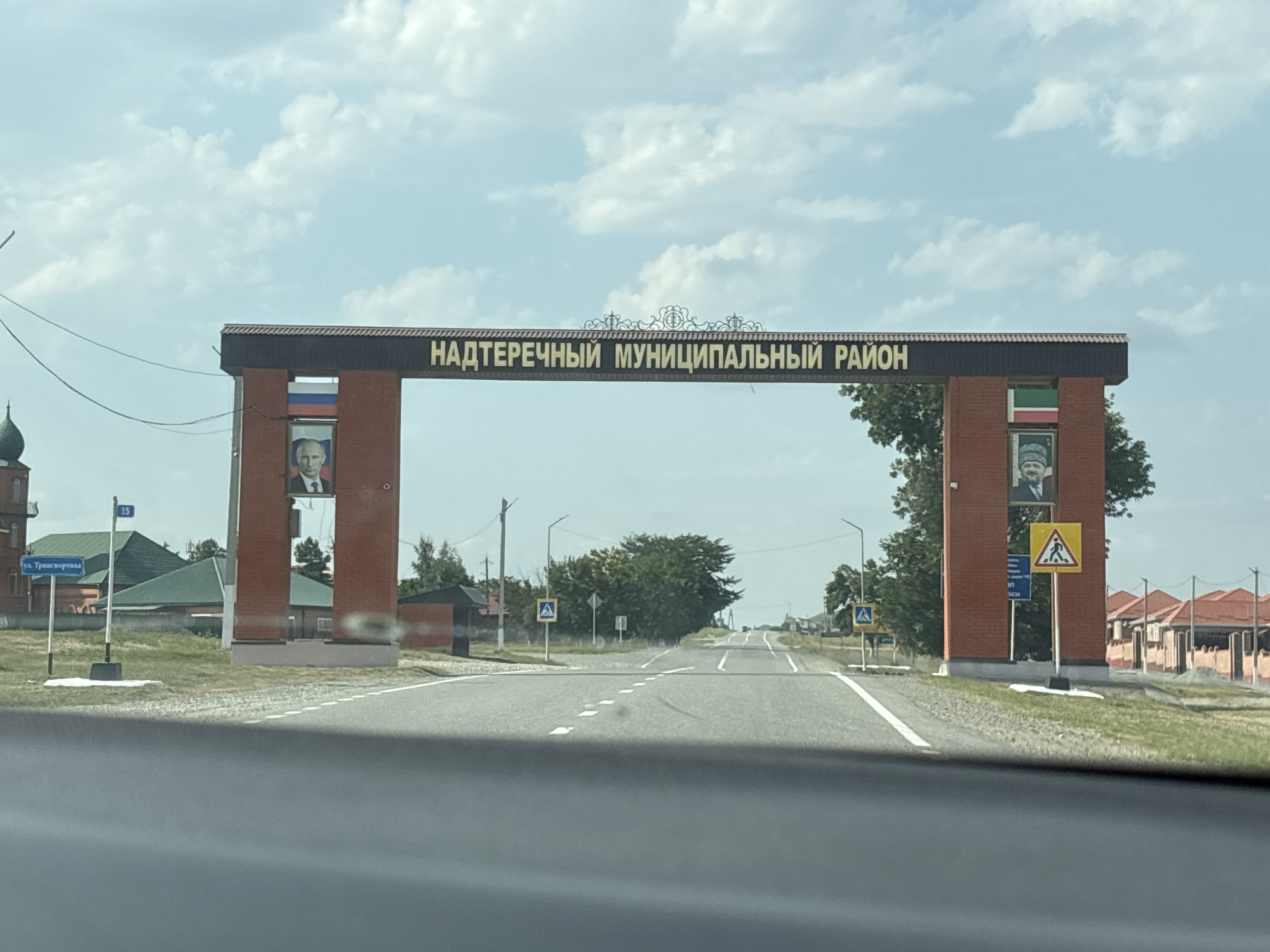 The image shows an arched gateway at the entrance to the Nadterechny Municipal District, located in Nagornoye, Russia. The gateway is made of red brick and features an inscription in gold letters that reads "НАДТЕРЕЧНЫЙ МУНИЦИПАЛЬНЫЙ РАЙОН" (Nadterechny Municipal District). On the left pillar, there is a portrait of a man wearing a suit. Above the portrait, a Russian flag is displayed. On the right pillar, there is a similar portrait of a man wearing a patterned headdress and a suit, along with a flag that appears to be green, white, and red. There are pedestrian crossing signs on both sides of the road leading through the gateway. The road is paved and marked with white lines. Houses with red roofs are visible on the right side of the image. The sky is partly cloudy. A street sign with "ул. Транспортная" (Transportnaya Street) is visible on the left side of the road.