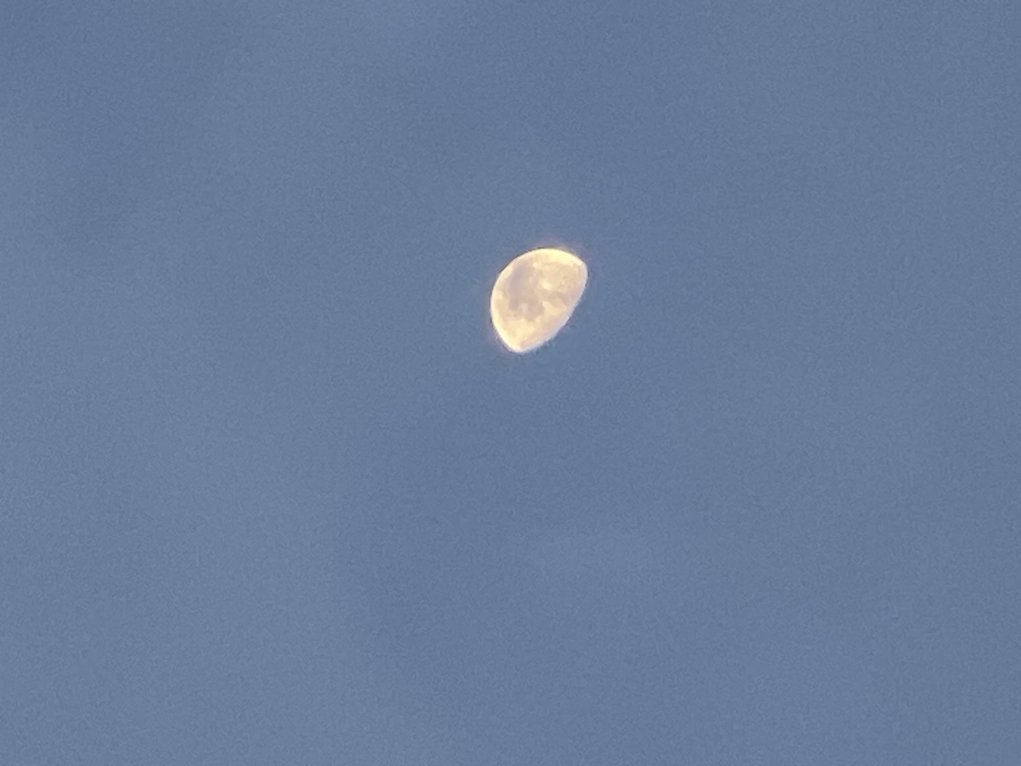 A crescent moon is visible against a clear blue sky. The moon is illuminated on its left side and appears to be partially obscured. The sky is a uniform shade of blue with no clouds or other celestial bodies visible. The image does not depict any elements related to the city of Yusuphana, Uzbekistan.