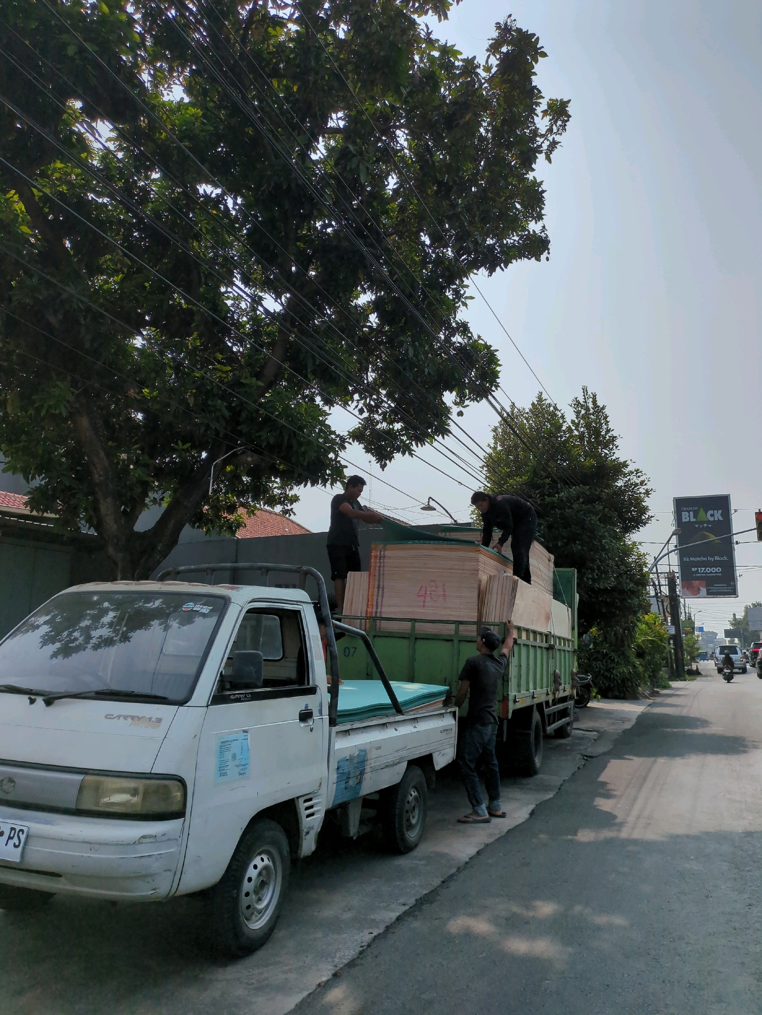 In Gentan, Indonesia, two men are seen unloading lumber from a green truck. A white pickup truck is parked alongside the green truck. The scene is captured on a street with trees and power lines overhead. A billboard is visible in the background.