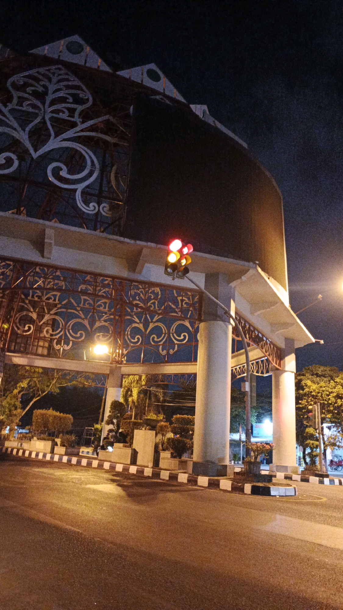 The image shows a traffic light at an intersection in the city of Surakarta, Indonesia. The traffic light is red and yellow, indicating a stop or caution. The intersection is paved with asphalt and has a striped curb. There are trees and decorative plants on the side of the road. In the background, there is a large, ornate structure, possibly part of a building or gate, with intricate metalwork. The sky is dark, suggesting it is nighttime. The scene appears to be quiet, with no visible people or vehicles actively in motion, except for a red motorcycle visible in the distance on the right side of the image. The lighting suggests artificial illumination from streetlights and the traffic signal itself.