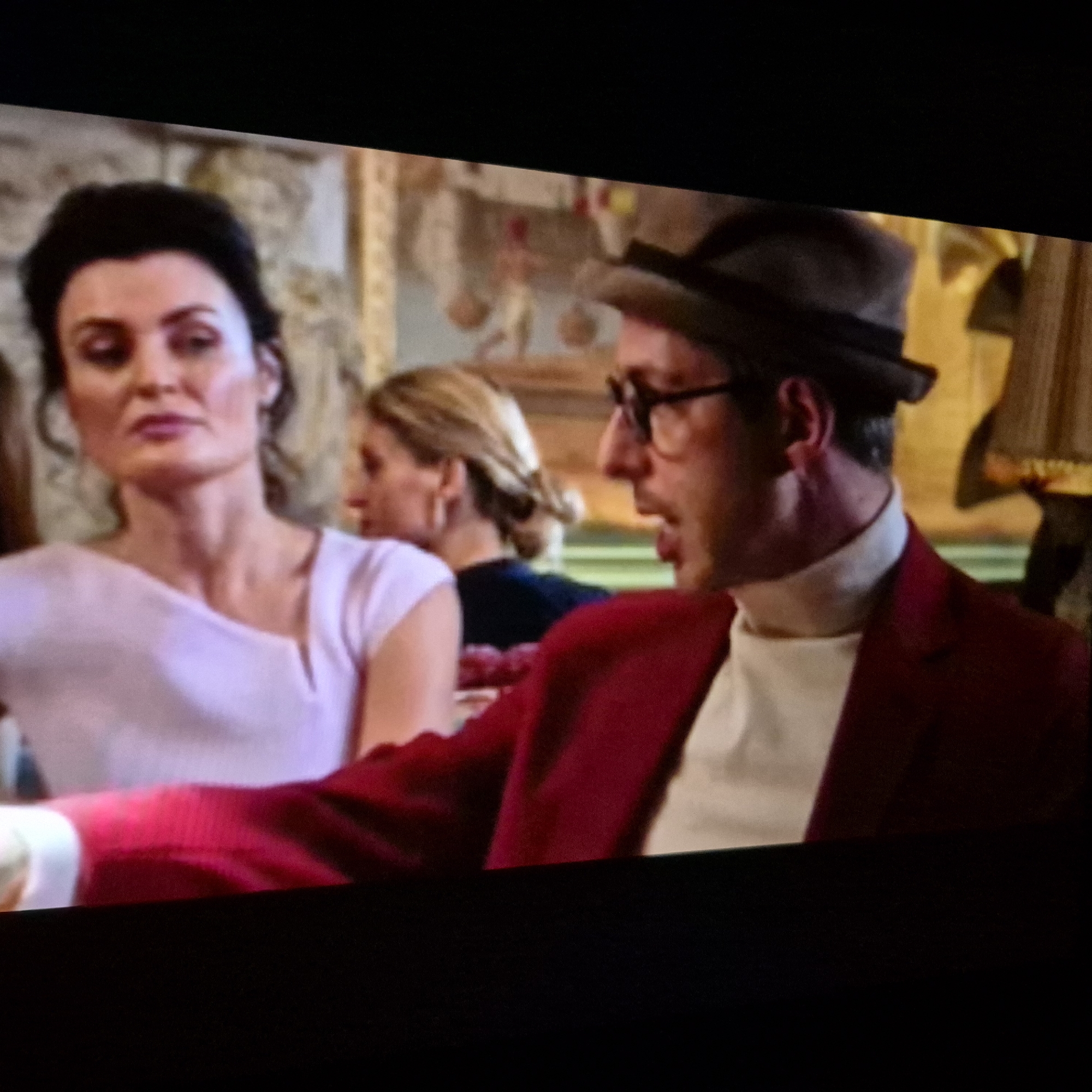 The image shows a medium shot of four people at an indoor event. On the left, a woman with dark hair pulled back is wearing a light pink off-the-shoulder top. Next to her, a woman with blonde hair in a bun is wearing a dark top. In the foreground on the right, a man wearing a brown hat, glasses, and a red jacket over a cream-colored turtleneck is speaking. Behind him, another person, likely a man based on clothing and hat, is visible. The background appears to be a banquet hall with ornate walls. The scene is captured from a slightly low angle, and the image has a grainy quality. The location in the city of Kępno, Poland is not specified by the image content.