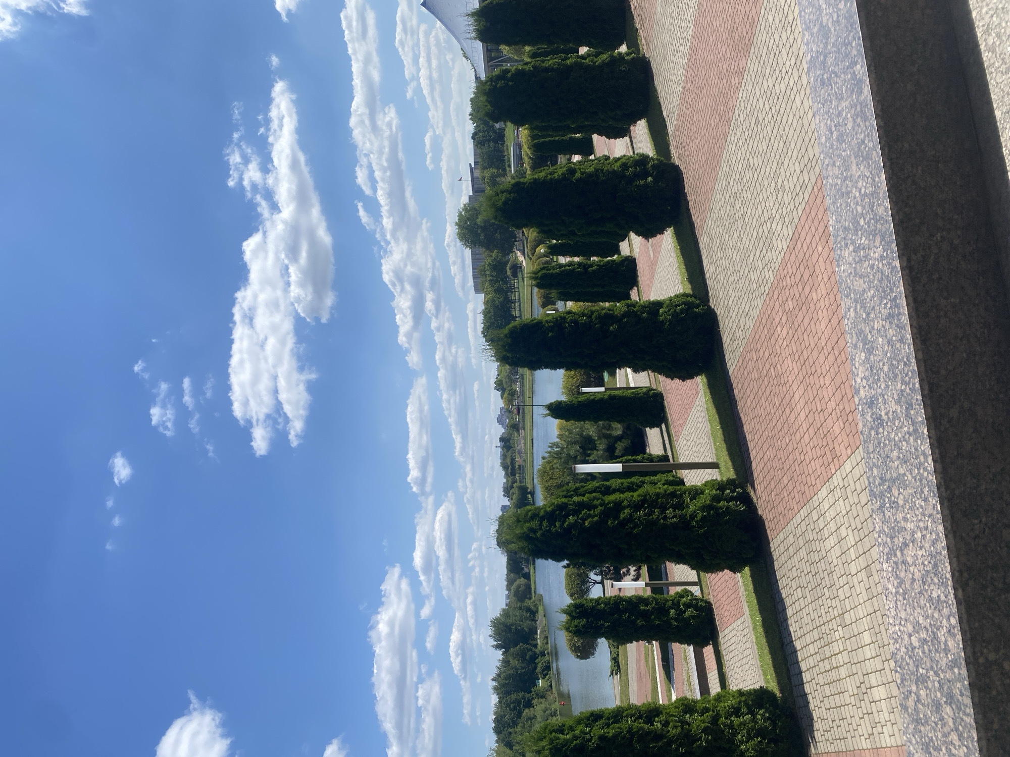 The image depicts a serene park scene with a body of water in the background, likely a lake or a large pond. Several well-maintained cypress trees are symmetrically planted in rows, creating a visual pathway. The ground is paved with a pattern of light and dark bricks. Beyond the trees and water, a treeline and some buildings are visible, suggesting an urban or suburban park setting. The sky is a clear blue with scattered white clouds. A granite-like structure is in the foreground, possibly a wall or a planter. The scene appears to be an outdoor recreational area within the city of Minsk, Belarus. No people or animals are present in the image.