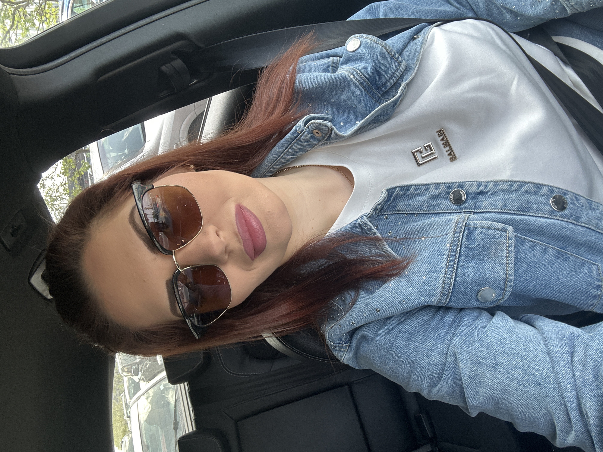 A woman with auburn hair and wearing sunglasses is sitting in a car. She is wearing a white shirt with a metallic "Balmain" logo and a light blue denim jacket adorned with rhinestones. The interior of the car is black. The location is not identifiable as Sofia, Bulgaria based on the visual information provided.