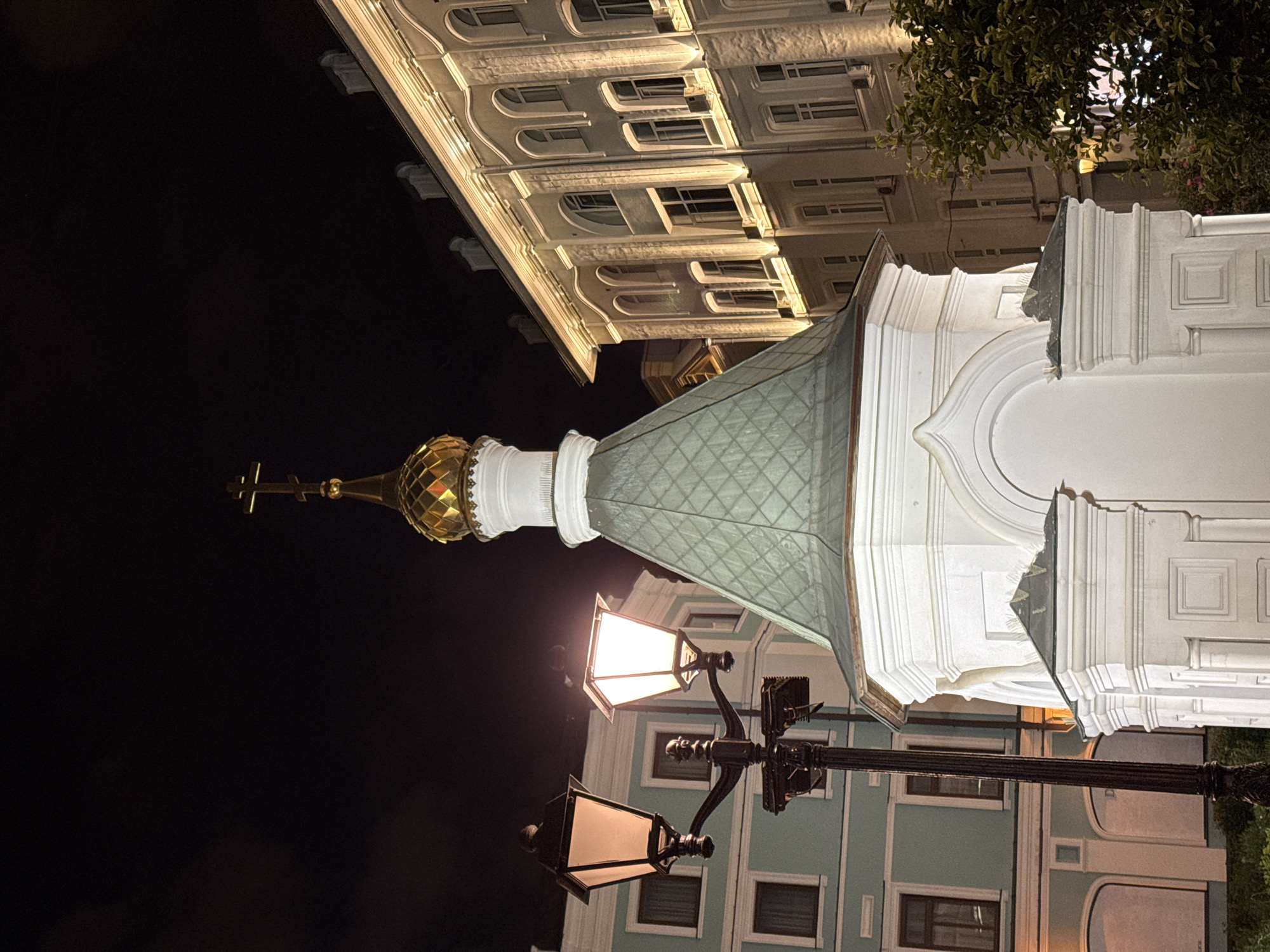 A low-angle, nighttime, outdoor, medium shot captures a white, ornate chapel with a green, patterned roof and a golden dome topped with a cross. The chapel is illuminated by external lighting. To the left of the chapel is a black lamppost with two lantern heads, one of which is lit. Behind the chapel and lamppost are two buildings. The building directly behind the lamppost is light blue with white trim and dark windows. The building to the right of the chapel is a light-colored, multi-story structure with arched windows and exterior lighting. The sky is dark and overcast. This scene is the Chapel of the Kazan Icon of the Mother of God located on Nikolskaya Street in Moscow, Russia.