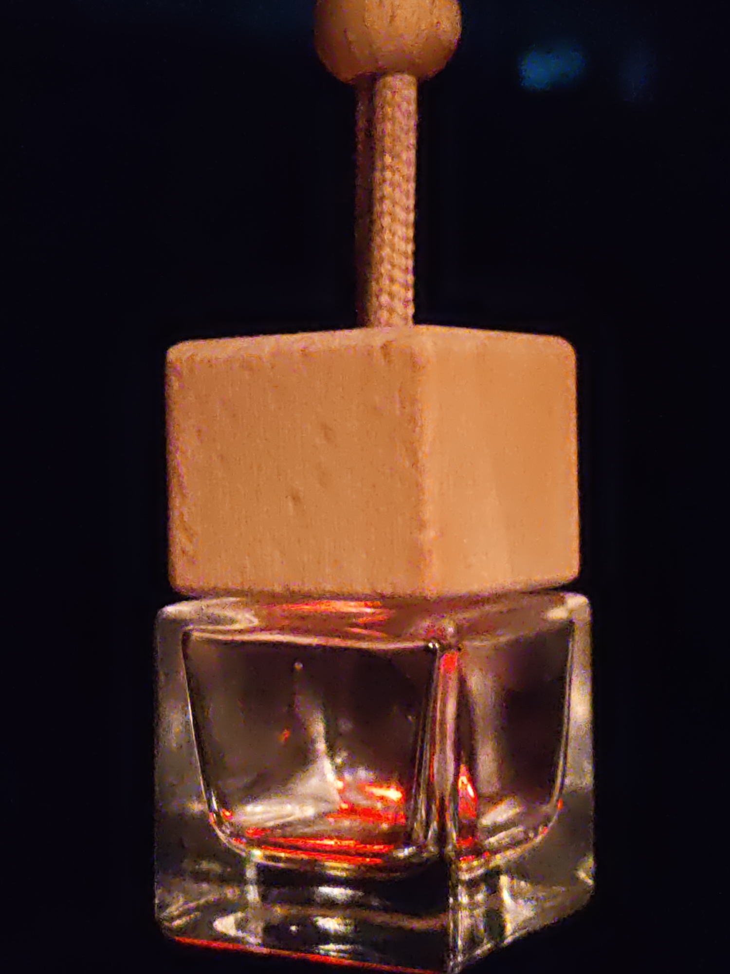 The image shows a close-up of a car air freshener. It is made of a wooden cube with a rope attached to a wooden bead at the top. The cube is suspended above a small glass bottle filled with a reddish liquid. The background is dark and out of focus, suggesting the air freshener is hanging. The image quality is somewhat grainy. There is no discernible location in the city of Olginskaya, Russia, or any people or actions depicted.