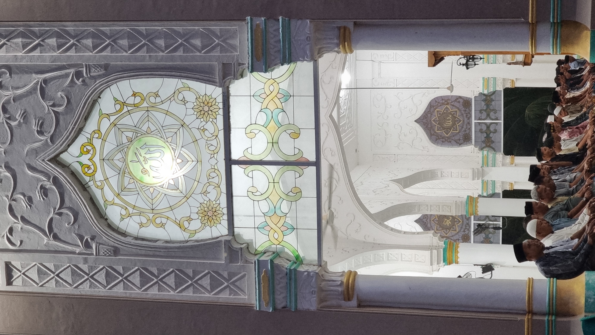 A group of men are seated on the floor in prayer inside a mosque. They are positioned in a line, facing forward, with their hands resting on their knees. The men are dressed in traditional Islamic attire, including long shirts and caps. The mosque's interior features white plastered walls with decorative arches and columns. A large stained-glass window with intricate geometric patterns and Arabic calligraphy is visible above the men. The scene appears to be in the city of Cotpuuk, Indonesia.