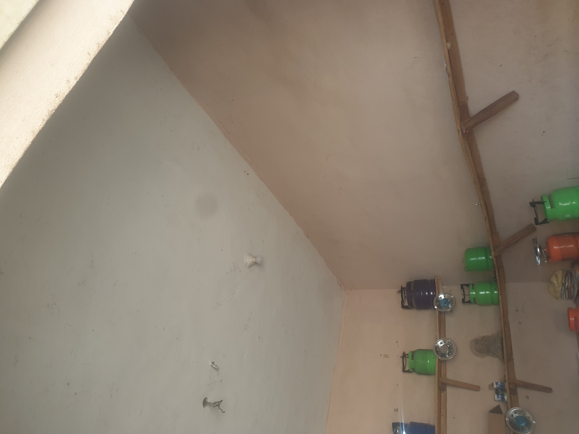 The image shows an interior view of what appears to be a storage area or a small shop in Jauro Yinu, Nigeria. The walls are painted in a light peach color, and the ceiling is a pale, textured white. Two wooden shelves are mounted on the walls, displaying various items.
On the upper shelf, from left to right, there are a blue gas cylinder, a lime green gas cylinder with a patterned texture, and a dark purple gas cylinder. Between the green and purple cylinders, there are two circular metallic objects, possibly hubcaps or reflectors.
On the lower shelf, there are two lime green gas cylinders and a stack of papers or documents. To the left of the lower shelf, a metallic circular object is visible, similar to those on the upper shelf. Further down, at the bottom of the image, more items are partially visible, including what looks like an orange gas cylinder and additional green gas cylinders. There are also some smaller containers and bottles on the shelves.
The ceiling has a light fixture with a bare bulb, and there are some minor imperfections and stains on its surface. Sunlight is entering the space from the upper right corner, casting a bright light and creating a distinct shadow line across the ceiling and the upper part of the wall. The overall impression is of a functional, possibly domestic or commercial storage space.