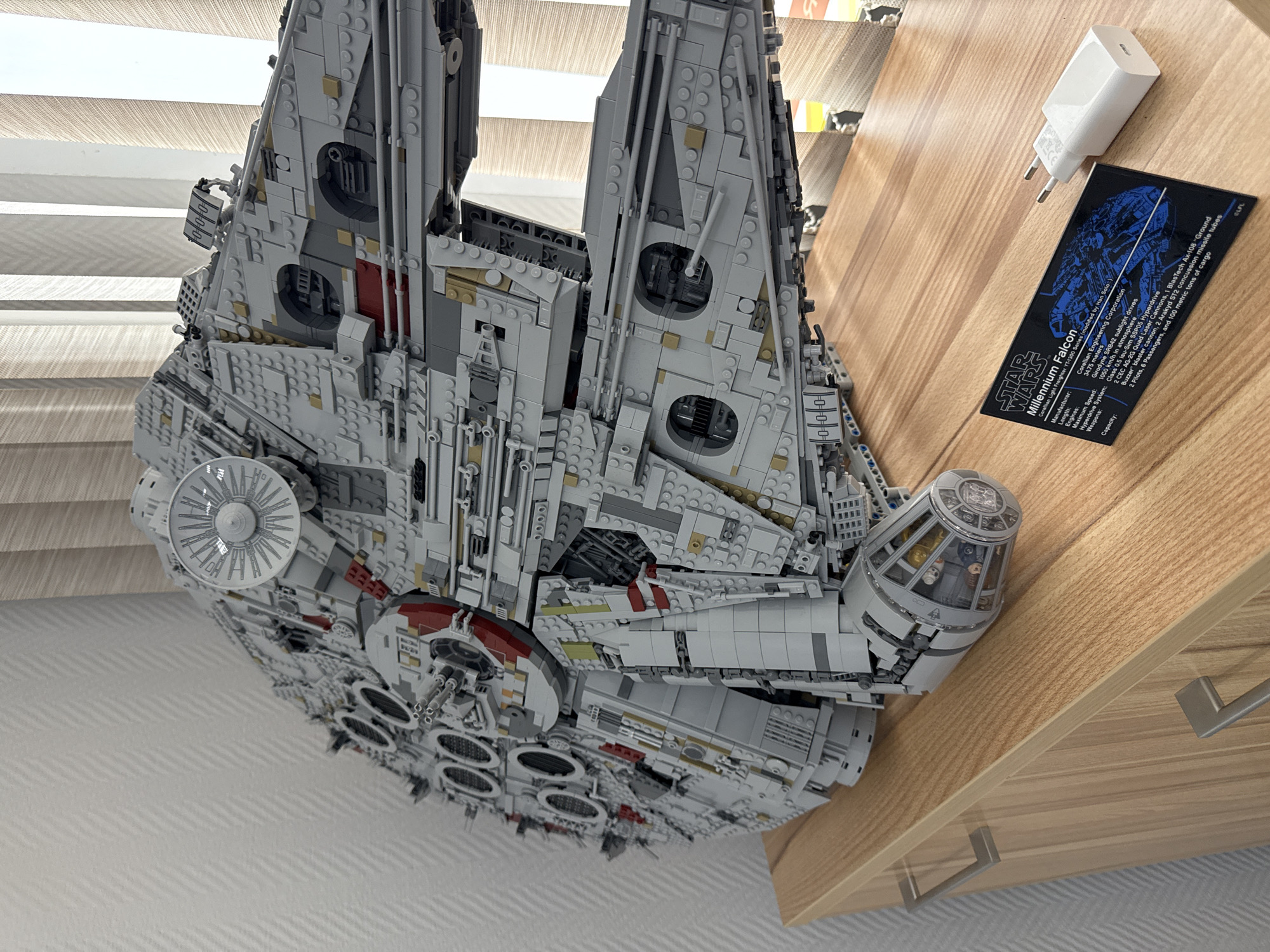 The image shows a Lego Millennium Falcon model resting on a wooden surface. Beside the model is a white USB power adapter and a black plaque displaying "Star Wars Millennium Falcon" along with technical specifications of the fictional spacecraft. The background includes a white blind and what appears to be a drawer or cabinet with metal handles. The image does not contain any information that places the subject in Krasnoyarsk, Russia, or any specific location.