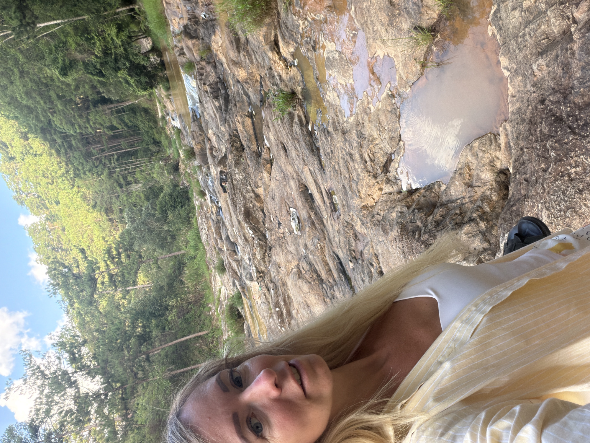 A Caucasian woman with blonde hair and blue eyes is taking a selfie in a natural environment. She is wearing a white top and a light yellow striped shirt. The background shows a rocky riverbed with shallow water and lush green trees on a hillside under a bright sky. The scene appears to be outdoors, possibly in a rural area of Păng Tiêng 1, Vietnam, given the vegetation and terrain. The woman's expression is neutral, and she is looking towards the camera.