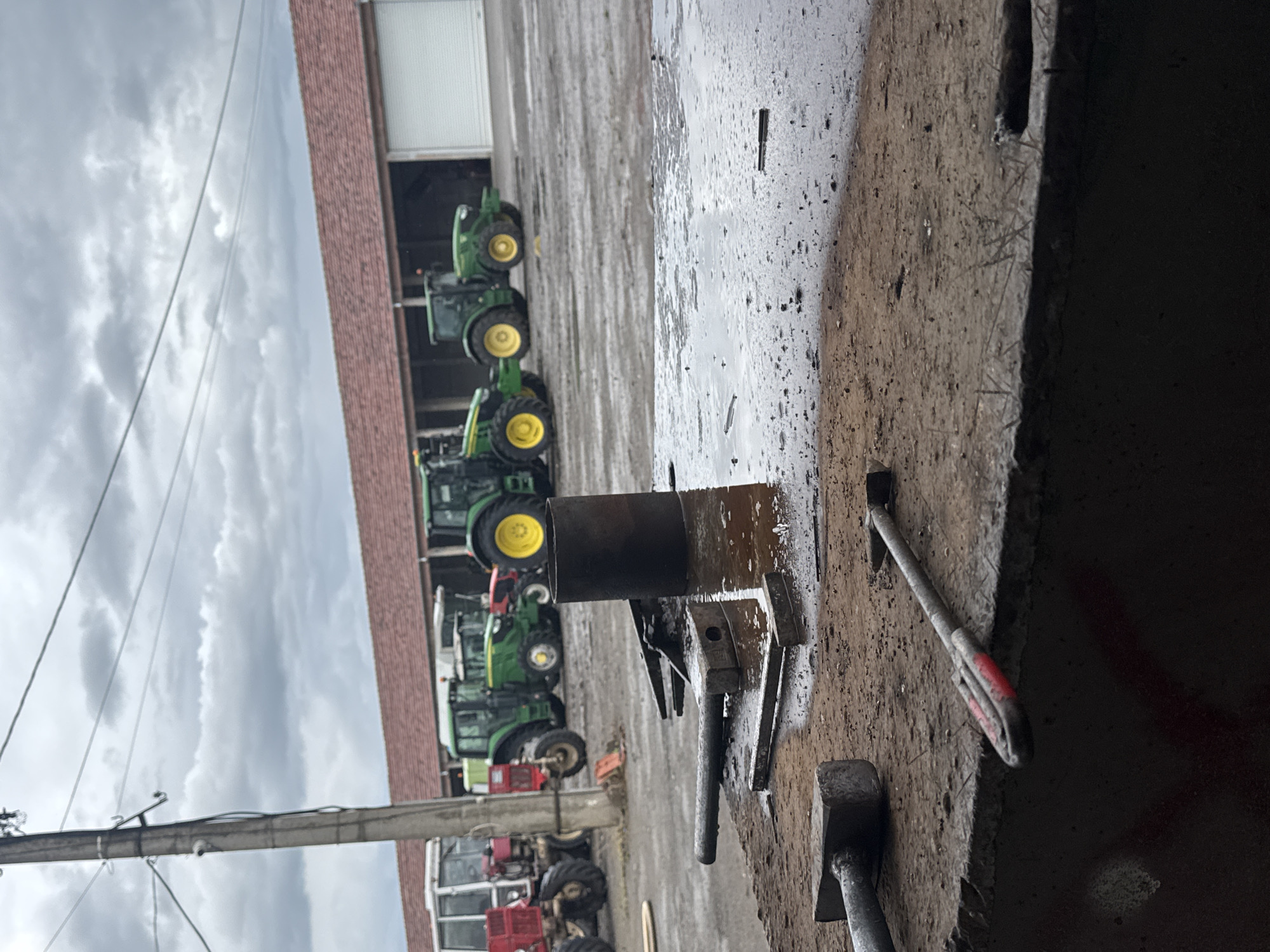 The image presents a scene in what appears to be an agricultural or industrial setting. A row of green and red tractors is visible in the background, parked in front of a long building with a garage door. In the foreground, a metal cylinder and various tools (including a hammer and possibly a wedge) rest on a concrete surface. The scene is somewhat weathered and appears to be outdoors on an overcast day. The location is specified as Sarnevo, Bulgaria.