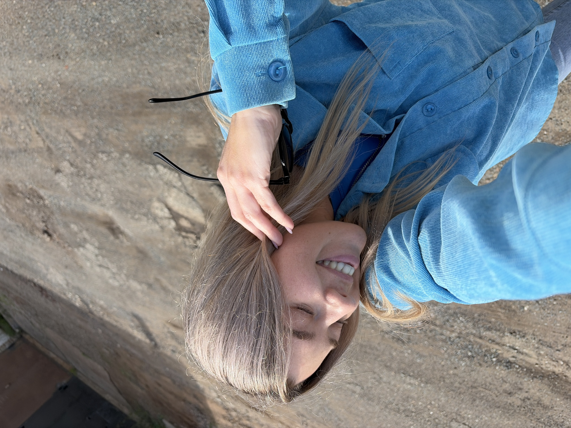 This high-angle, close-up image captures a young person, likely female, in Chaikovsky, Russia, from the chest up. The subject has long, light blonde hair and is positioned with their head tilted, eyes closed, and a wide, joyful smile. Their right hand is gently placed near their temple, touching their hair. In their left hand, a pair of black sunglasses is held close to their head.
The person is wearing a light blue corduroy jacket with visible buttons on the cuffs and front, indicating a casual and comfortable style. A darker blue garment or lining is visible underneath the jacket.
The background consists of a textured, grey-brown surface, resembling concrete or pavement, suggesting the person is lying down or leaning back on the ground outdoors. The bright, natural lighting indicates it is daytime, likely sunny or partly cloudy. The overall impression is one of relaxation, happiness, and contentment. No text is visible in the image.