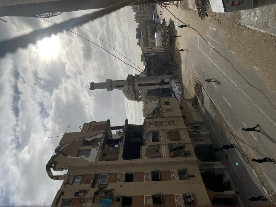 A cityscape reveals significant destruction, likely from conflict. Buildings are heavily damaged, some reduced to rubble. A mosque with a tall minaret stands amidst the destruction. A road is visible, with several people walking along it. The sky is cloudy and the sun is shining through the clouds. The overall scene suggests a post-conflict environment.