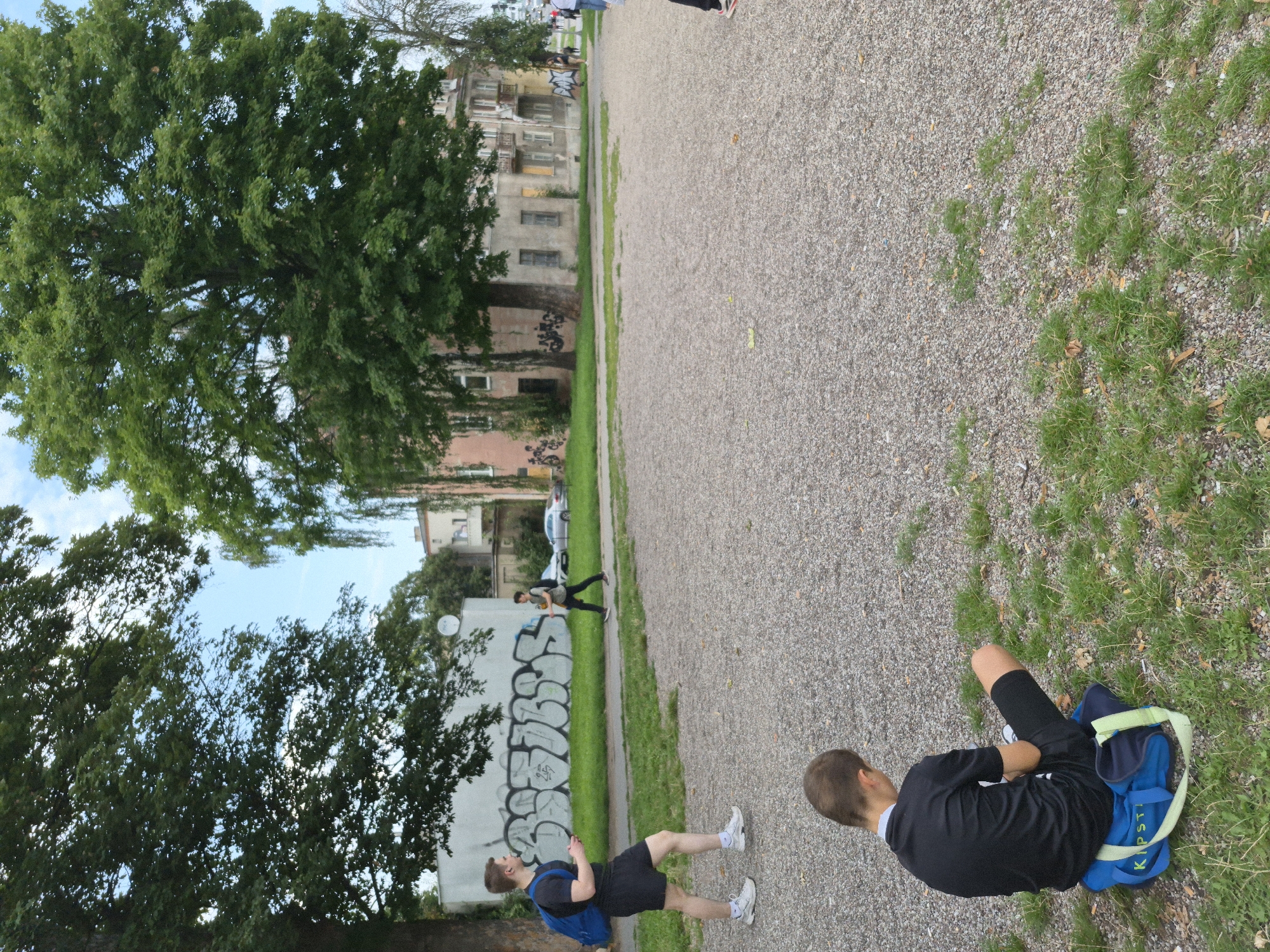 A boy in a black t-shirt and shorts sits on a blue and green sports bag on a patch of grass bordering a gravel area. He is facing away from the camera, looking down. In the background, two young men are walking across the gravel area. One, on the left, wears a black t-shirt, black shorts, a blue backpack, and white sneakers. He holds a phone in his right hand. The other, further to the right, wears a grey t-shirt, black pants, and a backpack. Behind them, a white wall with graffiti is visible, as well as trees and buildings, suggesting an urban environment. The scene appears to be in a park or public space within the city of Gdańsk, Poland, possibly near the historical Ołowianka island, given the architectural style of the buildings and the presence of a prominent tree line typical of the area.