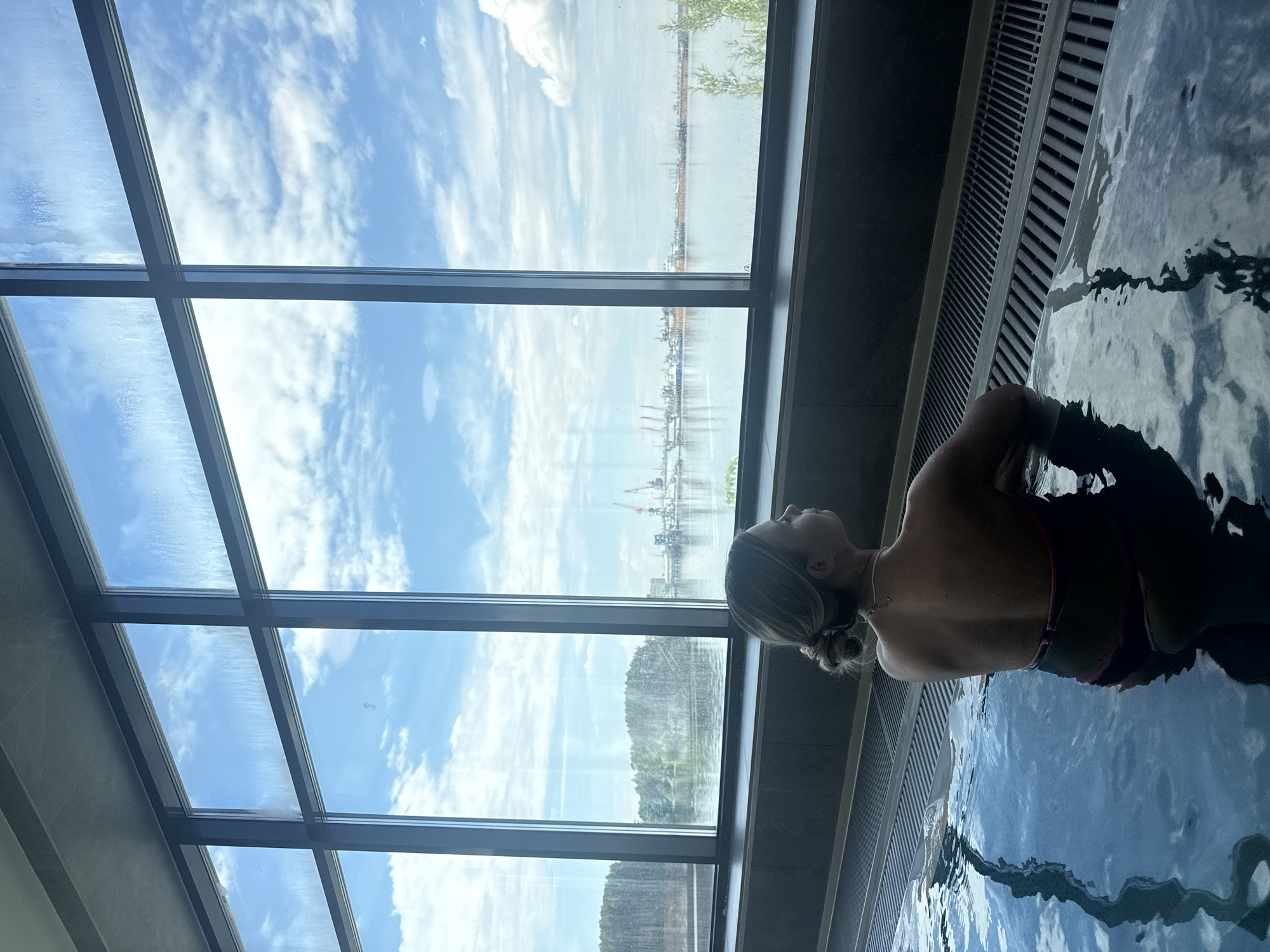 A woman is shown from the back and side view, with a glimpse of the right side of her face, while she is in the water. The surrounding area appears to be an indoor swimming pool, based on the presence of water, and the presence of a modern architectural design with expansive windows and architectural features. The woman is in the lower-right corner of the frame. She is looking out toward the windows on the left side of the frame. Outside the windows there is a water front and skyline. The windows are large and divided into sections. The skyline visible out the window includes buildings and infrastructure, appearing to possibly indicate a waterfront industrial area. The sky itself contains scattered clouds and a bright, daytime light. The overall setting evokes an indoor spa environment. The location is Chaikovsky, Russia, suggesting a modern or resort-style facility that offers swimming or water activities with a view of the local urban and natural landscape. The water surface displays ripples and light reflections, and the interior has a minimalist design.