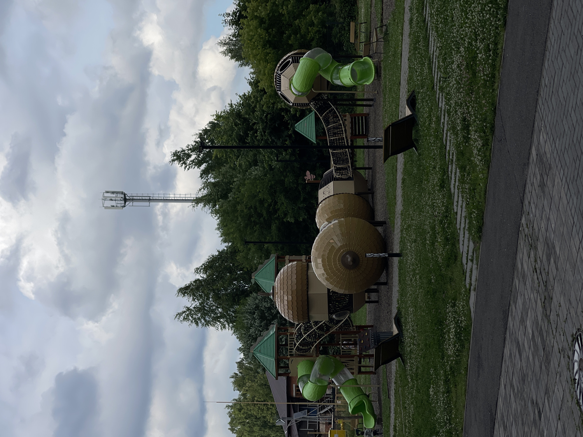 The image shows a modern playground with several large, whimsical structures designed to resemble acorns and spheres. The playground features two bright green spiral slides, a rope bridge connecting different platforms, and climbing walls. In the background, there are trees, a building, and a tall telecommunications tower under a cloudy sky. Two dark brown chaise lounges are positioned on the grassy area of the playground. The foreground shows a paved walkway with a cobblestone pattern. The overall scene depicts an outdoor recreational area.