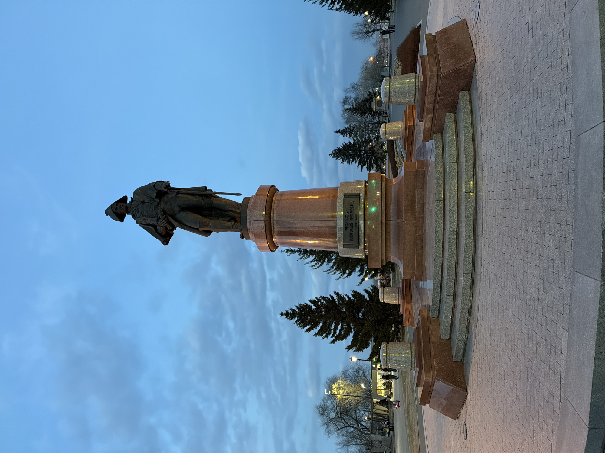 The image captures a bronze monument set against a blue, cloud-streaked sky. The monument depicts a figure in historical attire, possibly a soldier or explorer, mounted on a cylindrical plinth of polished red granite. The pedestal has a dark rectangular plaque mounted on it.
The monument is placed in a city park setting, with evergreen trees surrounding it and light posts illuminating the area. A paved walkway is prominent in the foreground, likely leading up to the statue. Other individuals are seen in the background walking along the pathway.
This is likely the monument to Andrey Dubensky, the founder of Krasnoyarsk.