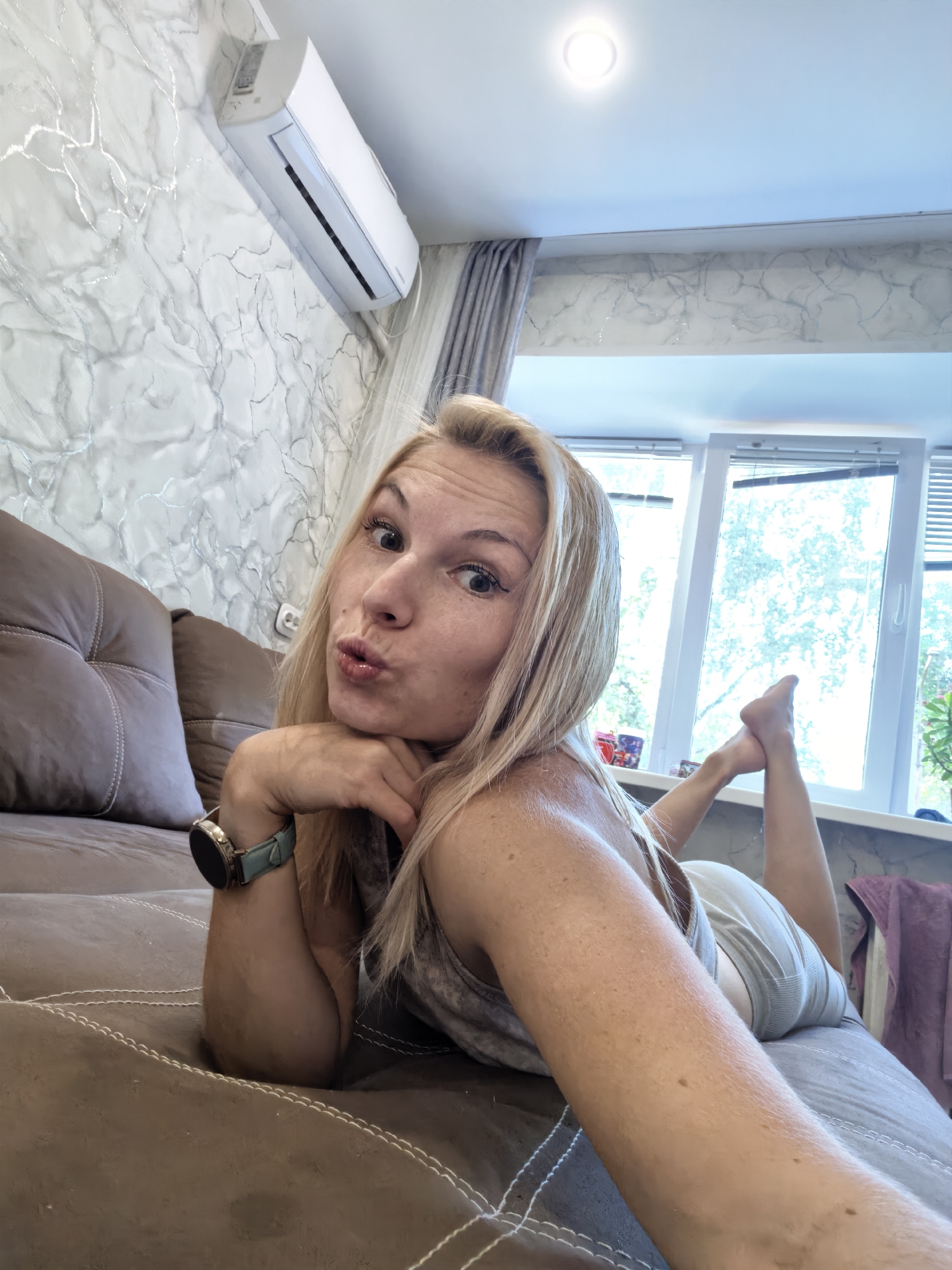 A blonde woman is lying on a brown couch in a room with light gray, textured walls featuring silver marble-like patterns. An air conditioning unit is mounted high on the wall. A large window with white blinds is visible behind her, and trees can be seen outside. The woman is looking at the camera with her head resting on her hand, her lips pursed. She is wearing a light-colored tank top and light shorts. A smartwatch with a light blue band is on her left wrist. The couch has visible stitching. The scene appears to be an indoor residential setting in the city of Sterlitamak, Russia.