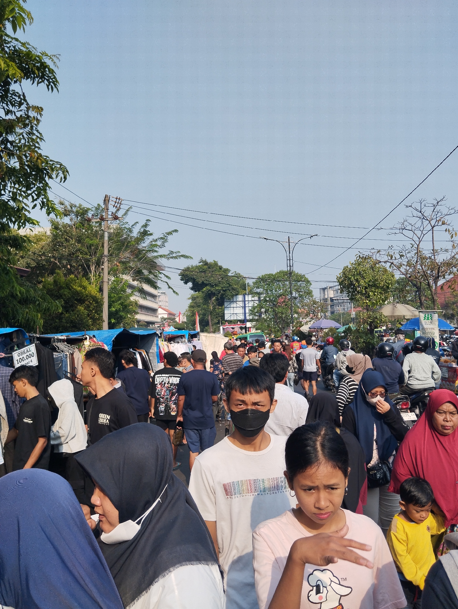 This is a medium shot, eye-level, outdoor photograph of a crowded street market scene in Surabaya, Indonesia, on a clear, sunny day. The foreground is dominated by several individuals. On the left, a person wearing a blue headscarf is partially visible. Next to them, a woman wearing a black hijab and a white shirt looks to her left. In the center, a young woman with dark hair, wearing a light pink t-shirt with a cartoon character, looks down with her hand raised, palm open. To her right, a young boy in a bright yellow t-shirt looks down. Further back, a man wearing a black mask and a white and black striped shirt looks forward.
Behind these foreground figures, the market stretches into the distance. Stalls with blue tarpaulin roofs are set up along the street, displaying various goods, primarily clothing on racks. People of all ages are browsing and walking through the crowded market. Some individuals are wearing masks. Motorcycles are also visible, with some people riding them. In the background, buildings and trees line the street, and overhead, power lines are strung between utility poles. The sky is a clear, light blue. A sign on a clothing rack in the left midground reads "OBRAL 100,000".