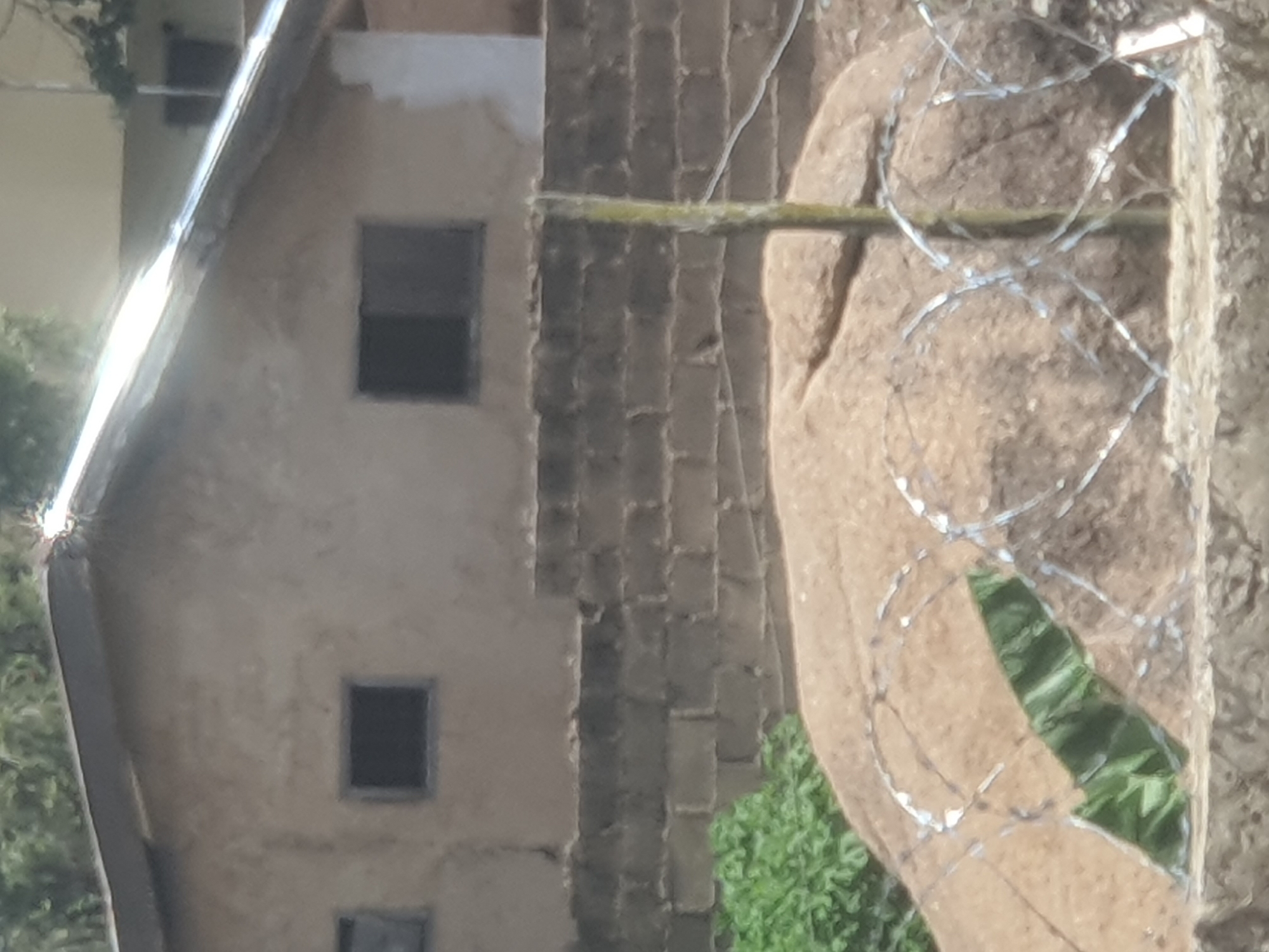 This is a medium-distance, slightly blurry, exterior photograph taken from a low angle looking up at a building and its surroundings. The building has a rough, light brown, stucco-like exterior with a corrugated metal roof that appears to be silver and shows signs of weathering. There are two dark rectangular windows visible on the upper part of the building. The building is situated behind a low wall constructed of rough, grey cinder blocks. In front of the cinder block wall, there is a large, amorphous mound of reddish-brown material, possibly sand or earth. This mound is partially covered by barbed wire fencing which is stretched across the foreground. A thin, weathered wooden or metal pole, stained yellow and green, stands vertically in front of the mound and to the right of the center of the image, running from the bottom edge upwards, passing through the barbed wire. To the left of the mound, some vibrant green foliage is visible, peeking out from behind the barbed wire. The overall setting appears to be an outdoor, possibly semi-rural or undeveloped area in the city of Jos, Nigeria, given the building style, materials, and vegetation. The lighting suggests it is daytime, with some bright spots on the roof indicating sunlight. No people or animals are present in the frame.