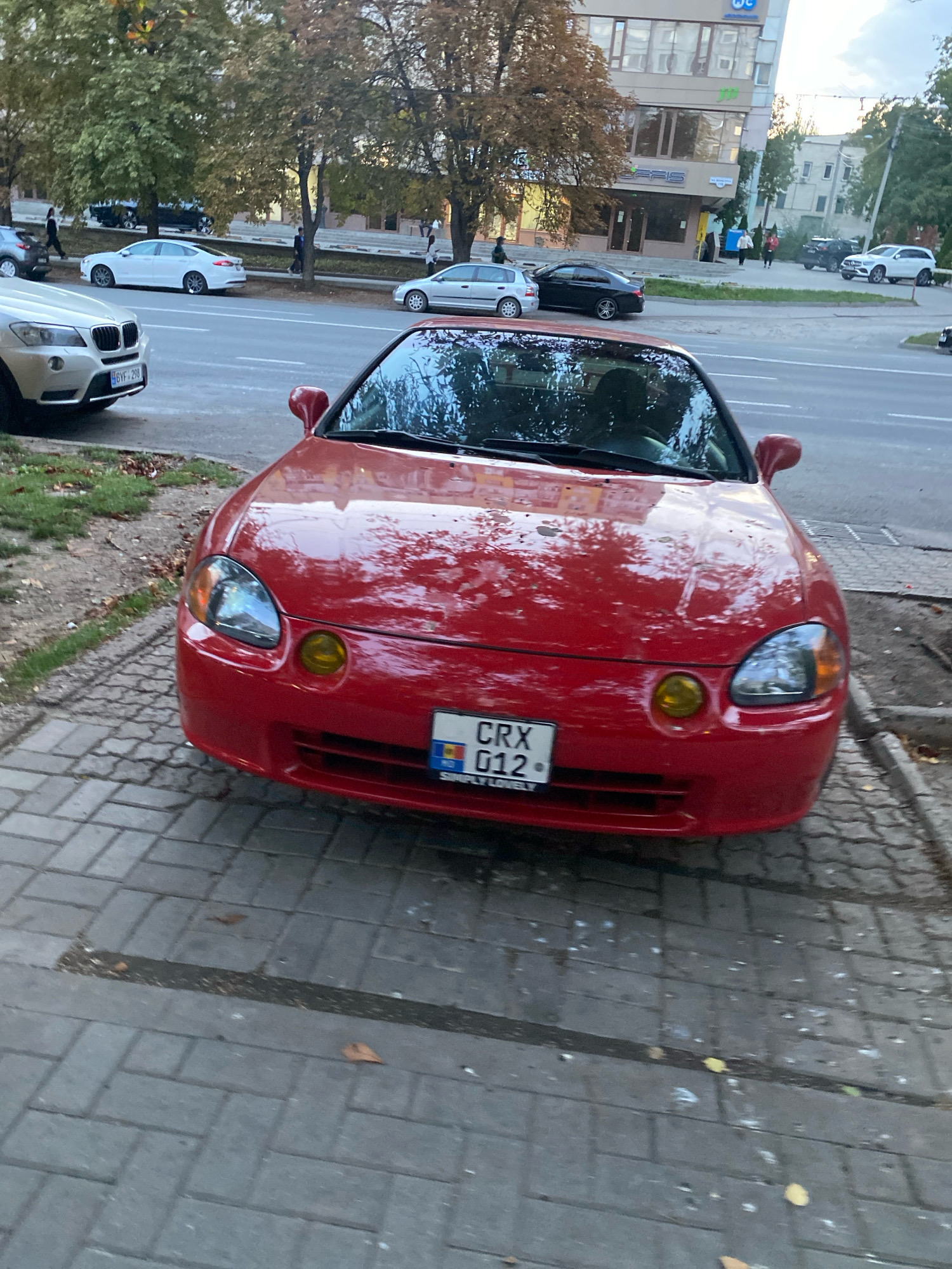 A red Honda CRX is parked on a sidewalk made of interlocking concrete pavers. The car has a license plate that reads "CRX 012" with the Moldovan flag on the left side. The street is visible behind the car, with other vehicles and pedestrians in the background. A building with several stories and windows is on the far side of the street. Trees with autumn foliage are also present. The scene appears to be in the city of Chișinău, Moldova, as indicated by the license plate.