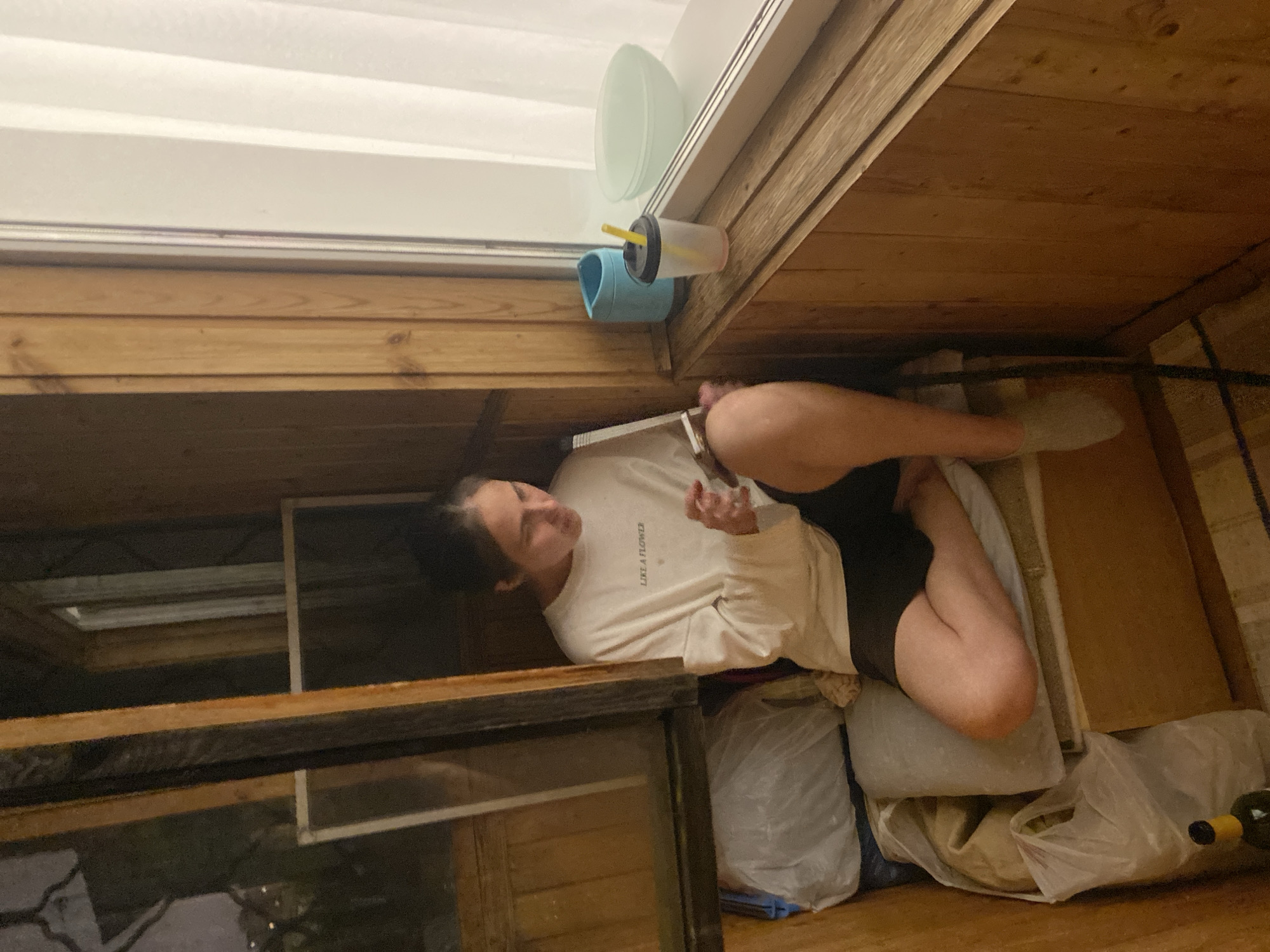 A young woman is captured in a low-angle, indoor shot, seemingly on a balcony or in a covered outdoor space with wooden plank walls. She is lying down, leaning against a stack of soft objects, possibly bags or cushions, and appears to be looking at her phone. She is wearing a long-sleeved white t-shirt with text that reads "LIKE A FLOWER" and dark shorts. Her legs are bent, and her right arm is extended, holding the phone.
Above her, on a wooden ledge, are several objects including a clear cup with a yellow straw, a blue holder for the cup, and a translucent pale green bowl. The background features a white horizontal surface, possibly a window or a wall, with horizontal lines suggesting blinds or panels. The overall setting suggests a casual, possibly cluttered, personal space. The lighting is somewhat dim, indicating it might be evening or an overcast day. The mood appears relaxed and informal. There are no obvious cues about the weather, but the presence of the cup and straw might suggest a beverage. The visible text on her shirt is the only discernible text in the image. The image is oriented sideways.