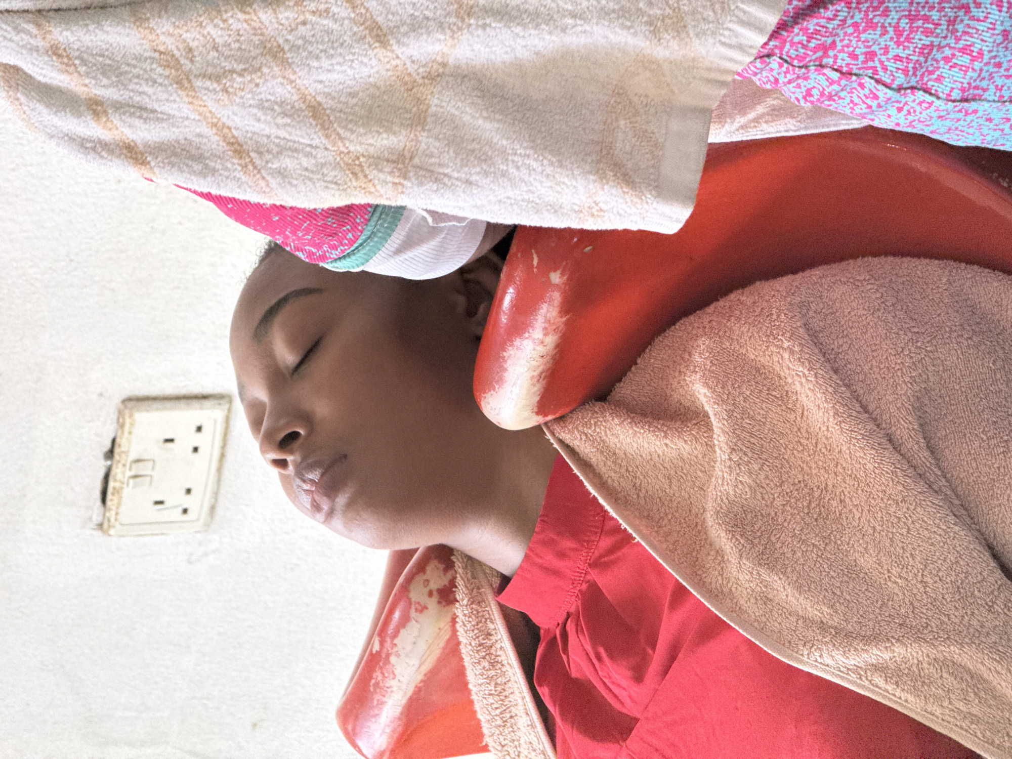 A close-up, upside-down, eye-level shot shows a dark-skinned person lying back in a red hair-washing basin with their eyes closed. Multiple towels in different colors (beige, white, and a blue/pink pattern) are draped around their head and the basin. The person is wearing a red shirt. A white electrical outlet with dark spots is visible on the white wall. The location is likely a hair salon in Abuja, Nigeria.