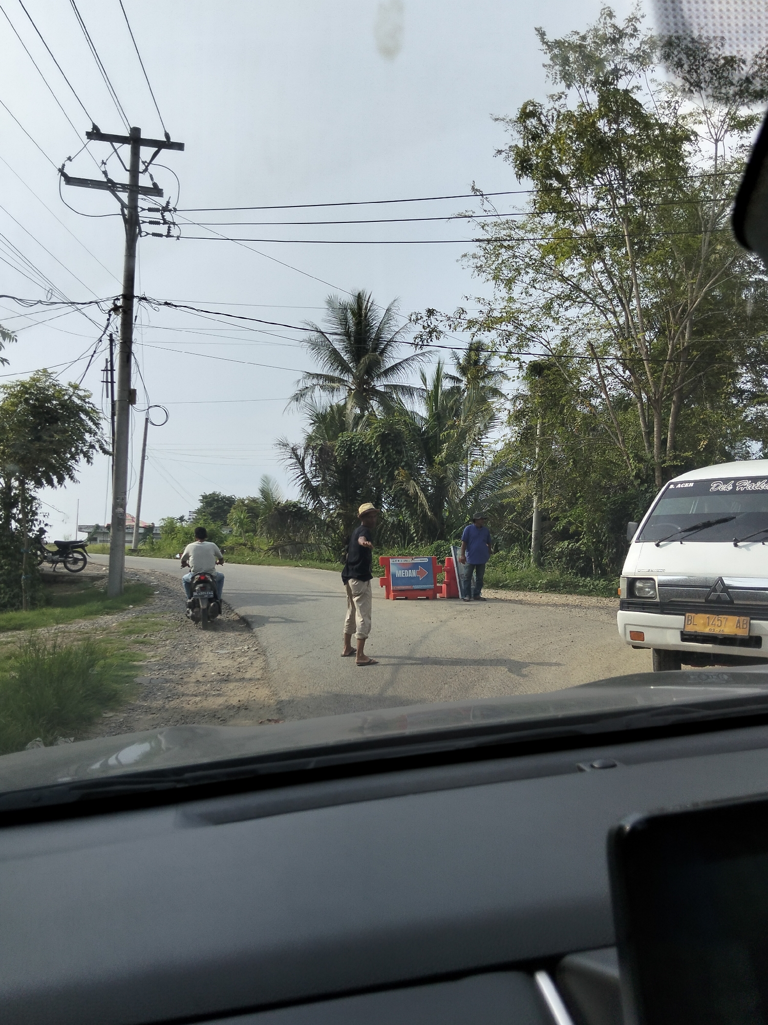 The image depicts a scene on a road in Blang Mee, Indonesia. In the foreground, the dashboard of a vehicle is visible, suggesting the photo was taken from inside. The road itself is paved and appears to be a two-lane street with a dirt shoulder on the left.
Several people are present. A man wearing a light-colored t-shirt and a helmet is riding a motorcycle, heading away from the camera. Further down the road, a man in a dark shirt and light-colored pants, wearing a straw hat, stands facing the camera. Another man, wearing a blue shirt, stands to the right of the road, near some traffic barriers. Two orange traffic barriers with white and blue signage are placed on the road, indicating a closure or redirection.
A white van with a license plate reading "BL 1457 AB" is parked on the right side of the road, facing away from the camera. A motorcycle is parked on the left side of the road near a utility pole.
The environment is characterized by lush green vegetation, including palm trees, and utility poles with numerous wires stretching across the sky. The sky is overcast with a light-colored haze. The overall impression is a somewhat rural or suburban setting on a cloudy day.
