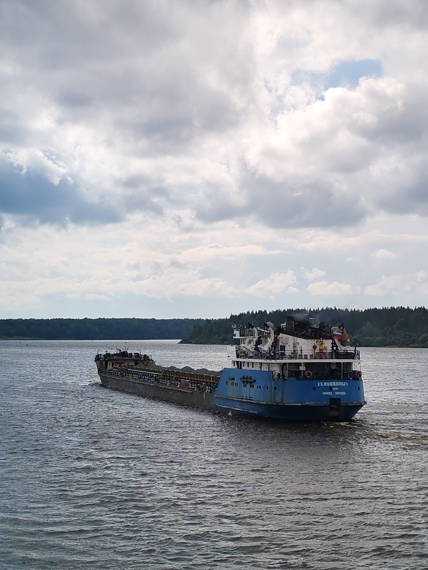 A blue and gray barge carrying cargo is moving through a wide, calm river. The barge has a multi-level superstructure with cranes and equipment visible. A Russian flag is displayed on the superstructure. The river is surrounded by a dense, dark green forest that stretches to the horizon under a cloudy sky. The water has a rippled texture from the movement of the barge. The scene appears to be in the vicinity of Даргун, Russia.