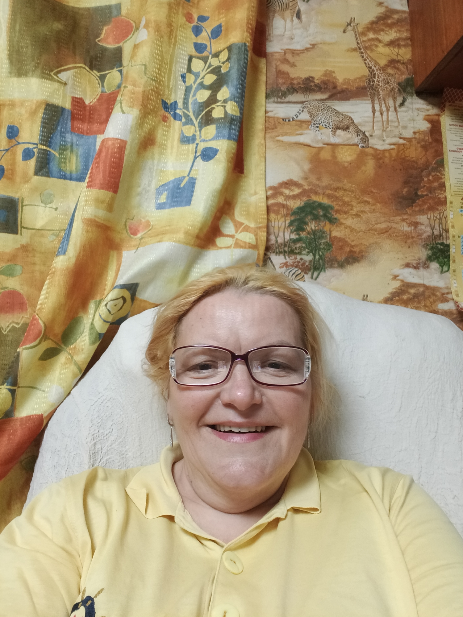 A medium shot depicts a fair-skinned, middle-aged woman wearing glasses and a yellow, button-down shirt, smiling broadly. She is seated against a white, textured pillow. The background is composed of patterned curtains featuring abstract shapes in orange, yellow, blue, and white on the left, and a wall decorated with an African savanna scene including zebras, a giraffe, and a leopard on the right. The city location is Unknown, Unknown.