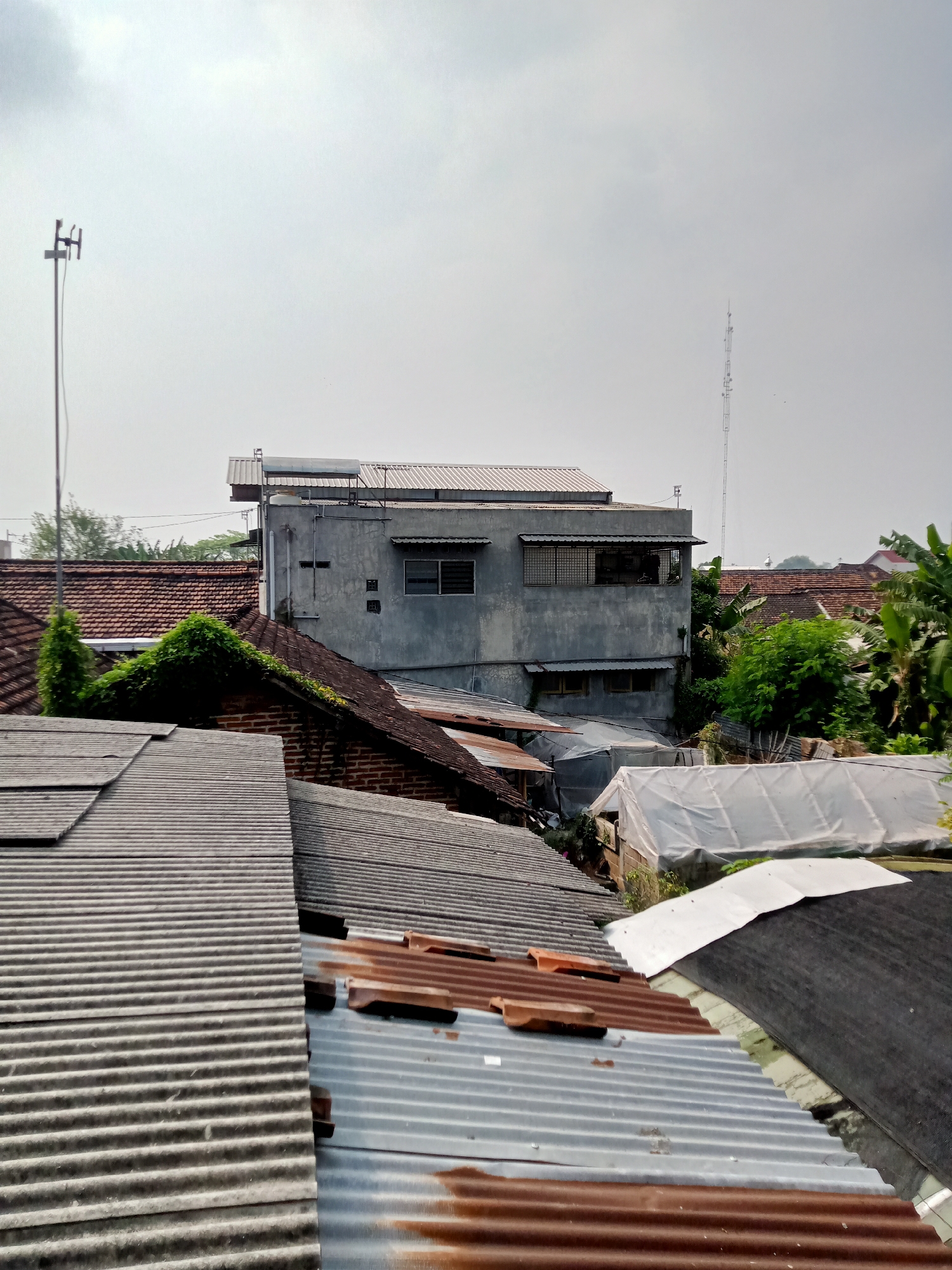 The image shows an aerial view of rooftops in the city of Simo, Indonesia. The foreground is dominated by various corrugated metal and tiled roofs, some appearing aged and rusted. In the mid-ground, a grey, multi-story building stands out with visible windows and what appears to be a solar water heater on its roof. Adjacent to this building are more rooftops, including structures covered in white sheeting, suggesting greenhouses or temporary shelters. Lush green trees and foliage are interspersed among the buildings, indicating a mix of urban and natural elements. In the background, a tall communication tower rises against a hazy, overcast sky. The overall scene depicts a dense residential or mixed-use area with a utilitarian aesthetic. No people are visible in the image.