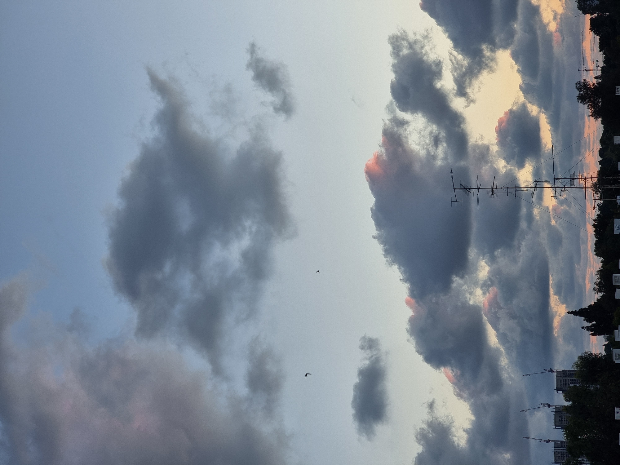The image shows a cloudy sky in Ramat Gan, Israel. The sky is filled with cumulonimbus clouds, varying in shades of gray and blue. The setting sun casts a faint orange glow on the horizon. In the background, there is a cityscape with buildings and construction cranes visible. An antenna is present. Birds are flying in the sky.