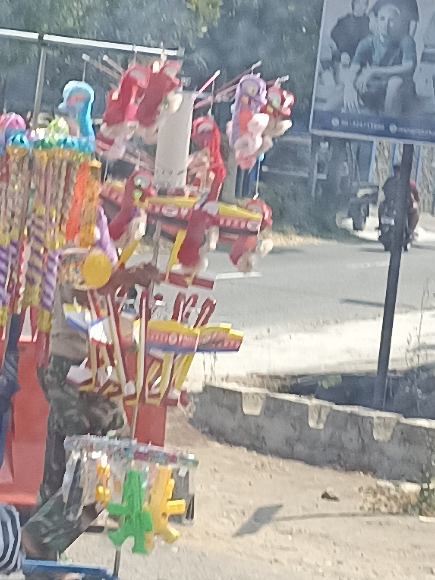 The image depicts a street vendor in Pulisen, Indonesia, selling an assortment of colorful novelty items. The vendor, partially visible and wearing camouflage clothing, is positioned in front of a stall displaying various toys and decorations. Hanging from the stall are bright red and pink stuffed toys, along with other items featuring yellow and purple striped patterns. In the foreground, a rack holds several small, plastic toys, including a green cactus-shaped item and a yellow star-shaped item. Behind the stall, a road is visible, with a few vehicles in the background. A large poster with images of people is also present on the right side of the frame, and a dark pole stands near the poster. The overall scene appears to be an outdoor market or roadside stall setting.